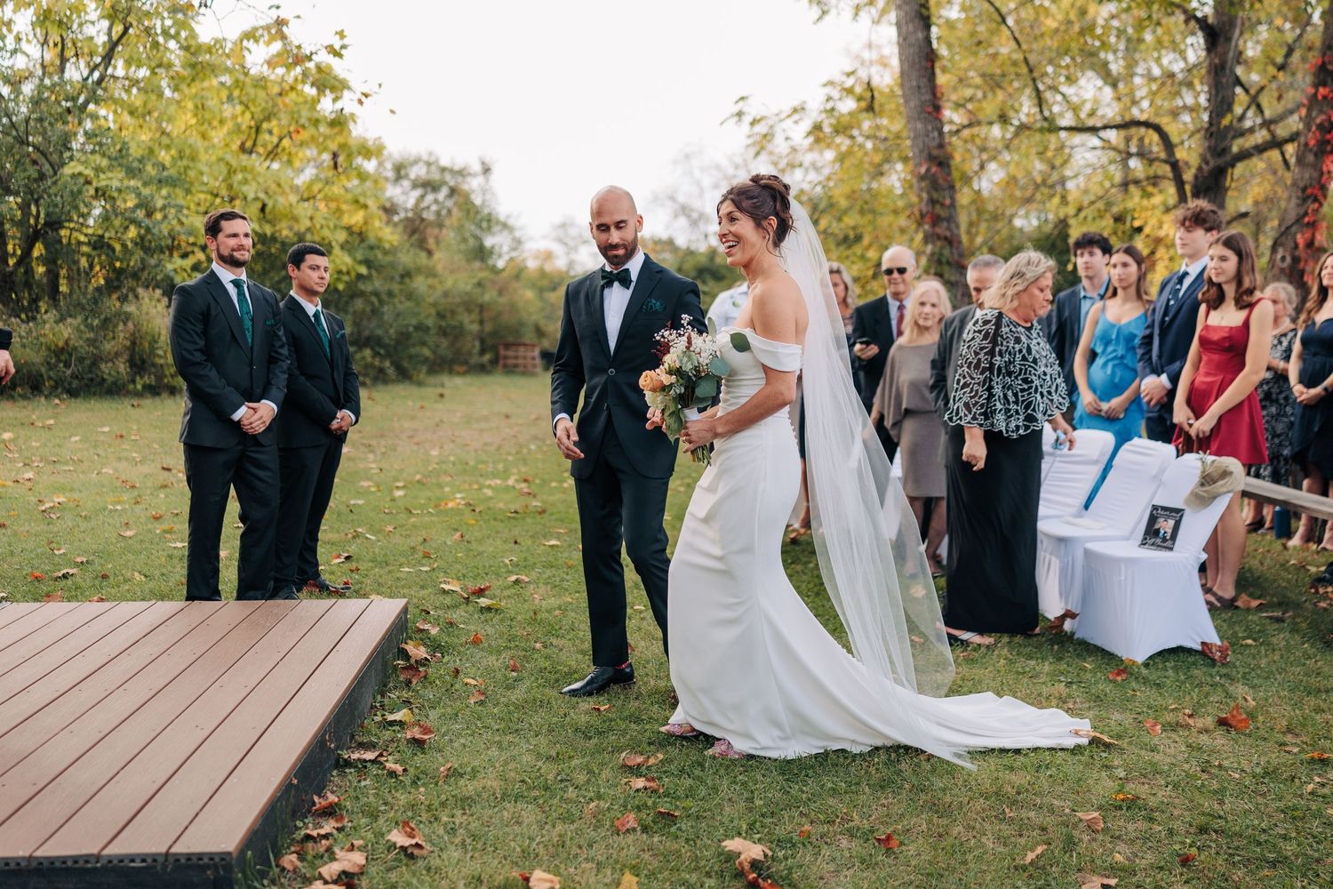 Outdoor wedding ceremony with guests seated on white chairs surrounded by fall foliage.