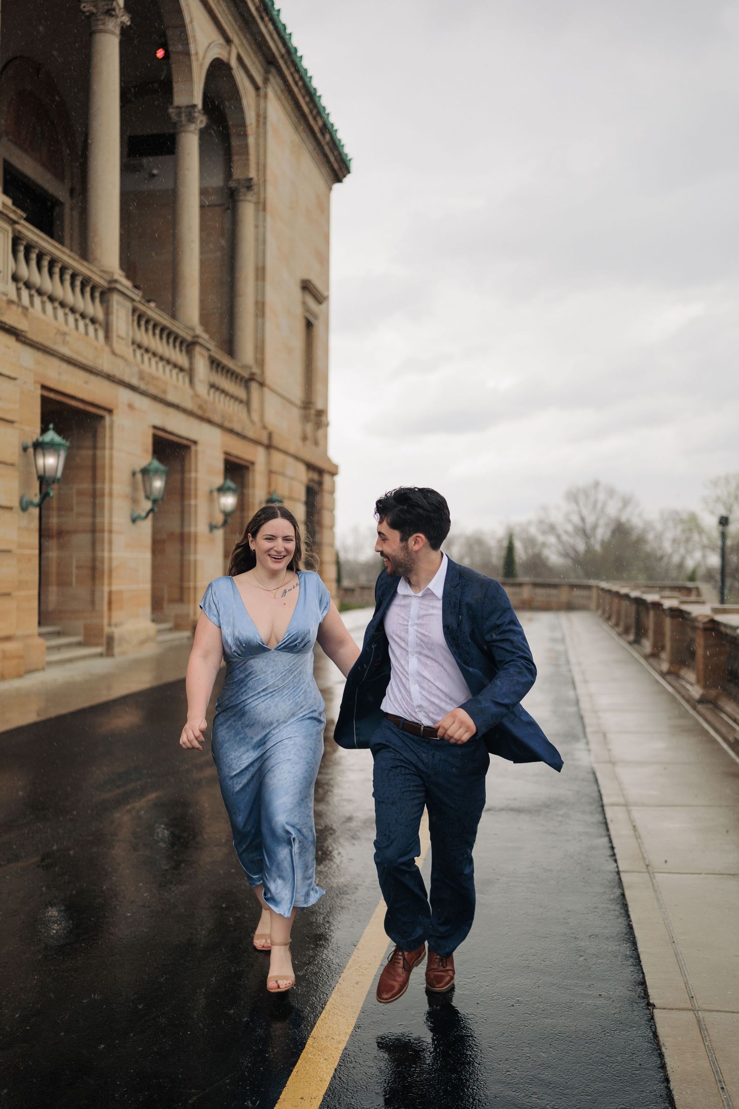 Couple laughing and running hand-in-hand on a wet sidewalk beside a stone building on a cloudy day.