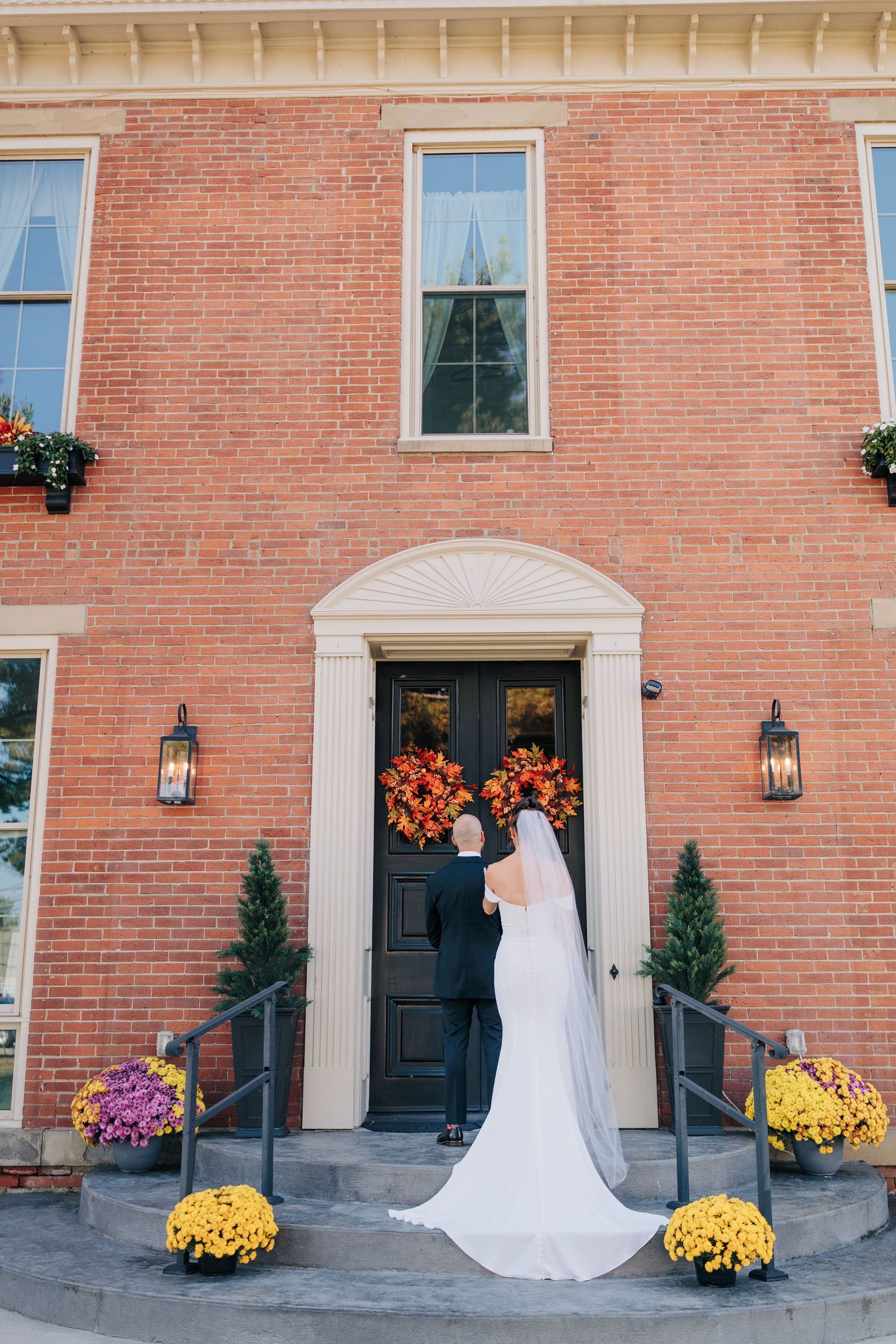 A white wedding gown flowing down steps of a brick building with fall mums and lanterns.