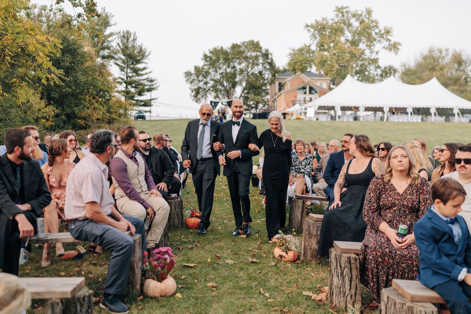 Wedding guests gather outdoors on a grassy lawn during an autumn celebration with fall foliage in the background.