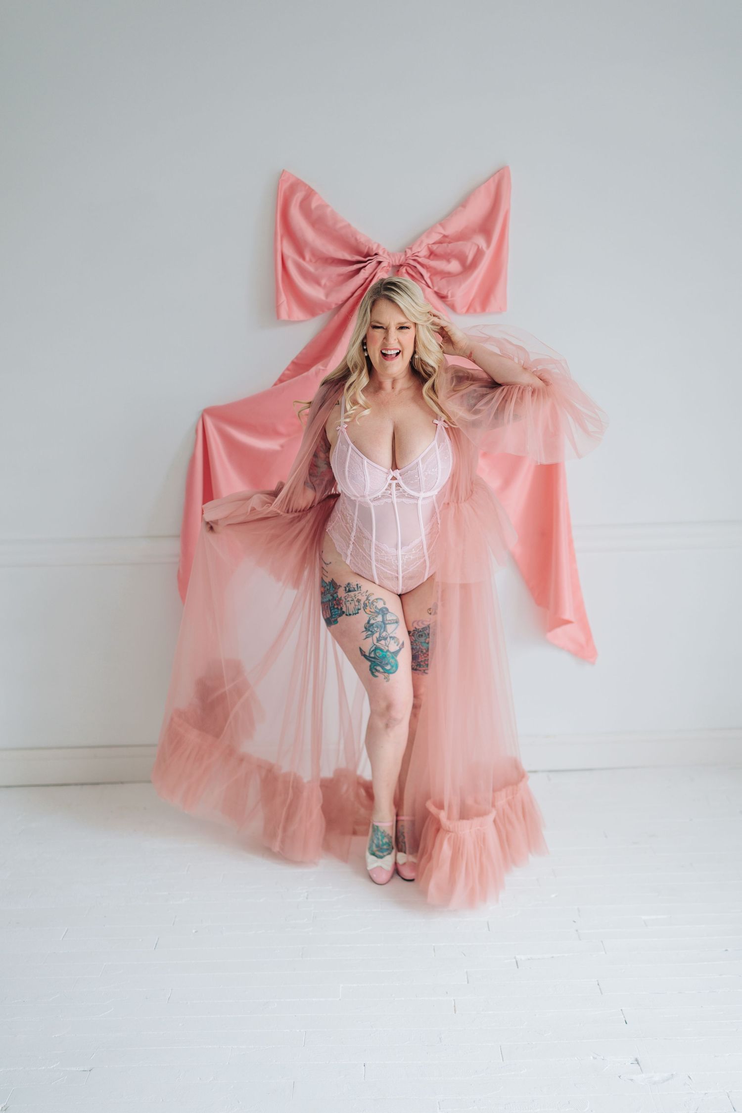 Boudoir photo with pink bow backdrop and sheer pink robe against white wall.