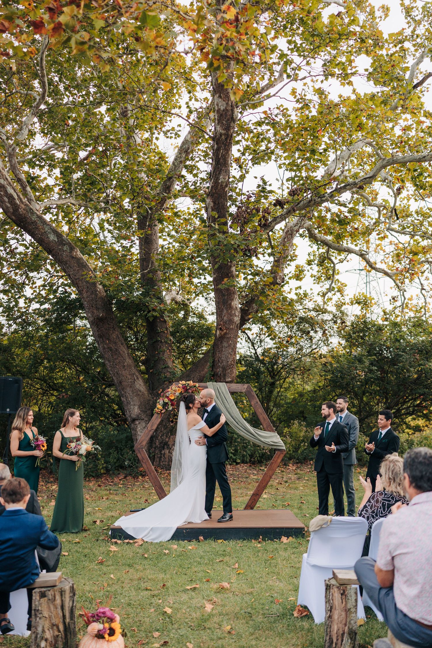 Wedding ceremony guests watching couple exchange vows under autumn trees.