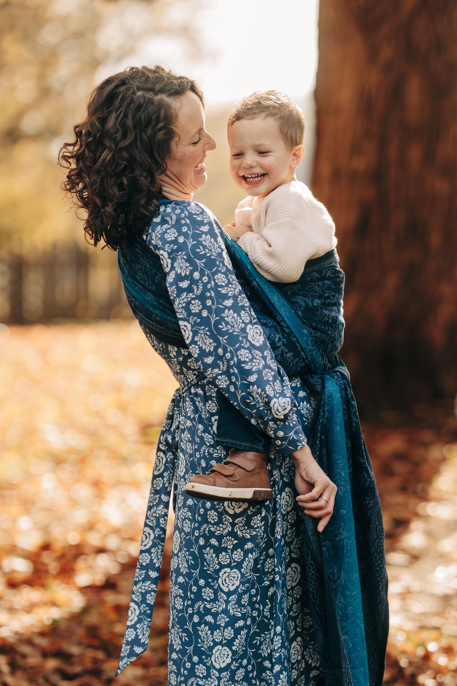 A mother and child share tender moments in a blue floral dress against an autumn backdrop of fallen leaves.