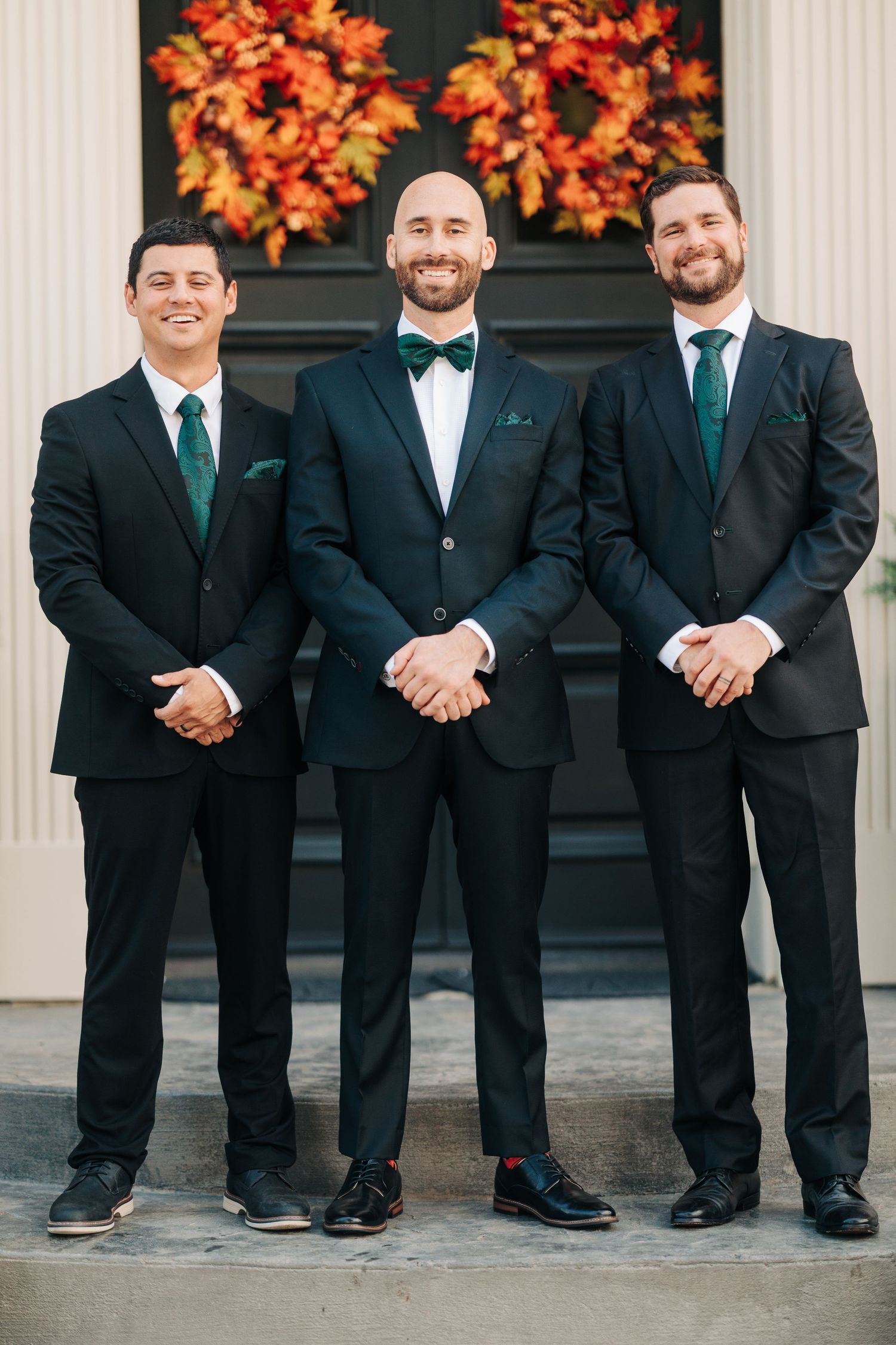 A line of groomsmen in black suits and emerald green ties stand together against a backdrop with orange fall foliage.