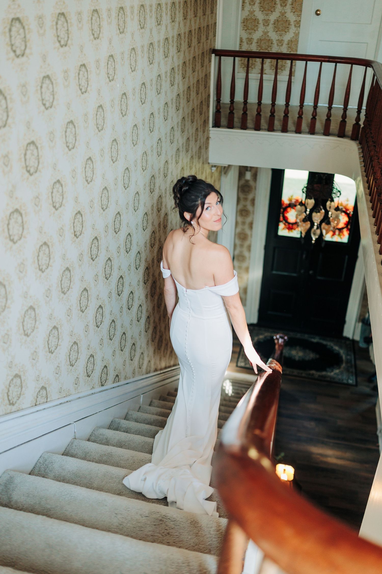 A bride in a white off-shoulder gown stands on carpeted stairs against vintage floral wallpaper in a historic home.