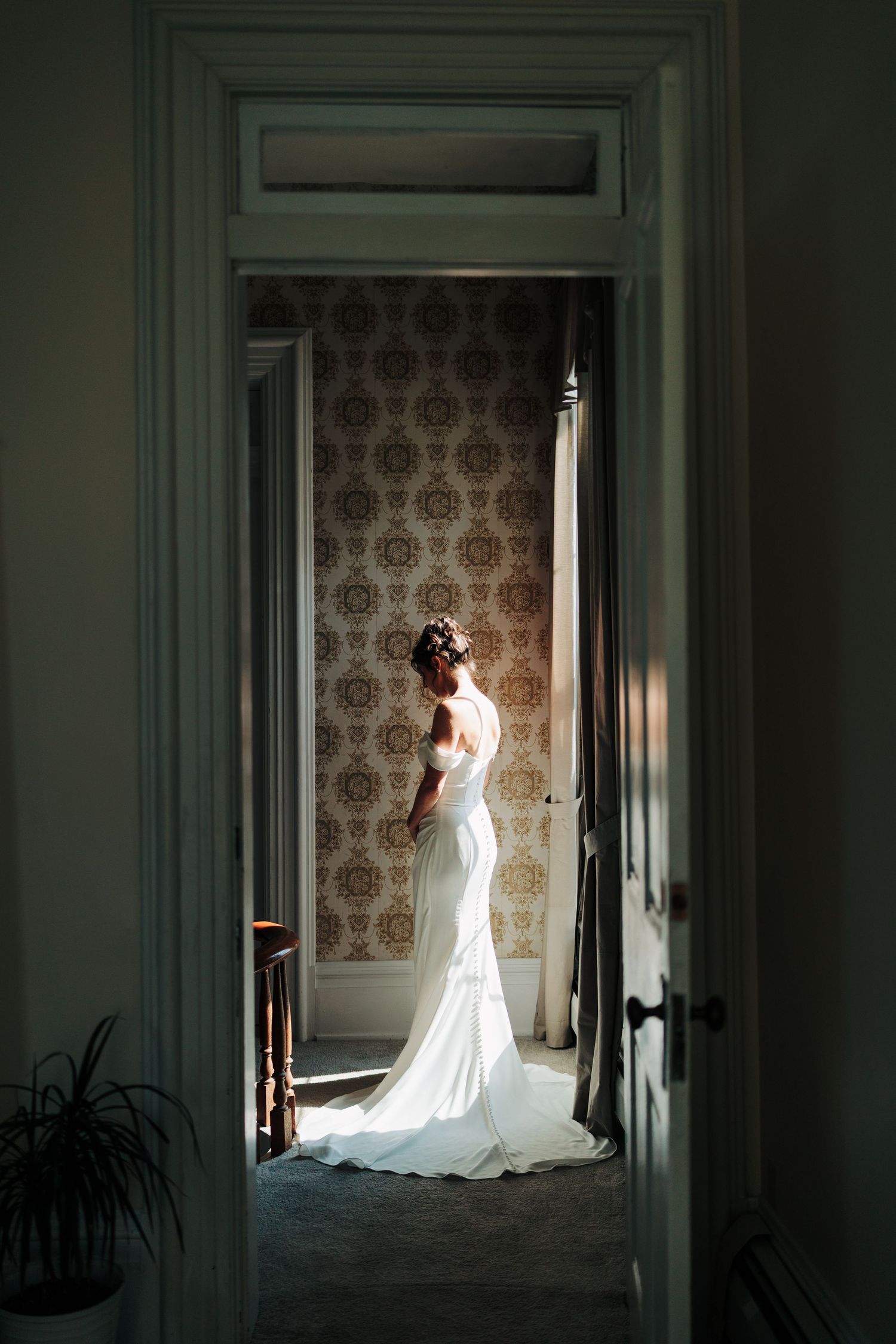 An elegant wedding dress silhouette framed in a doorway against vintage wallpaper.