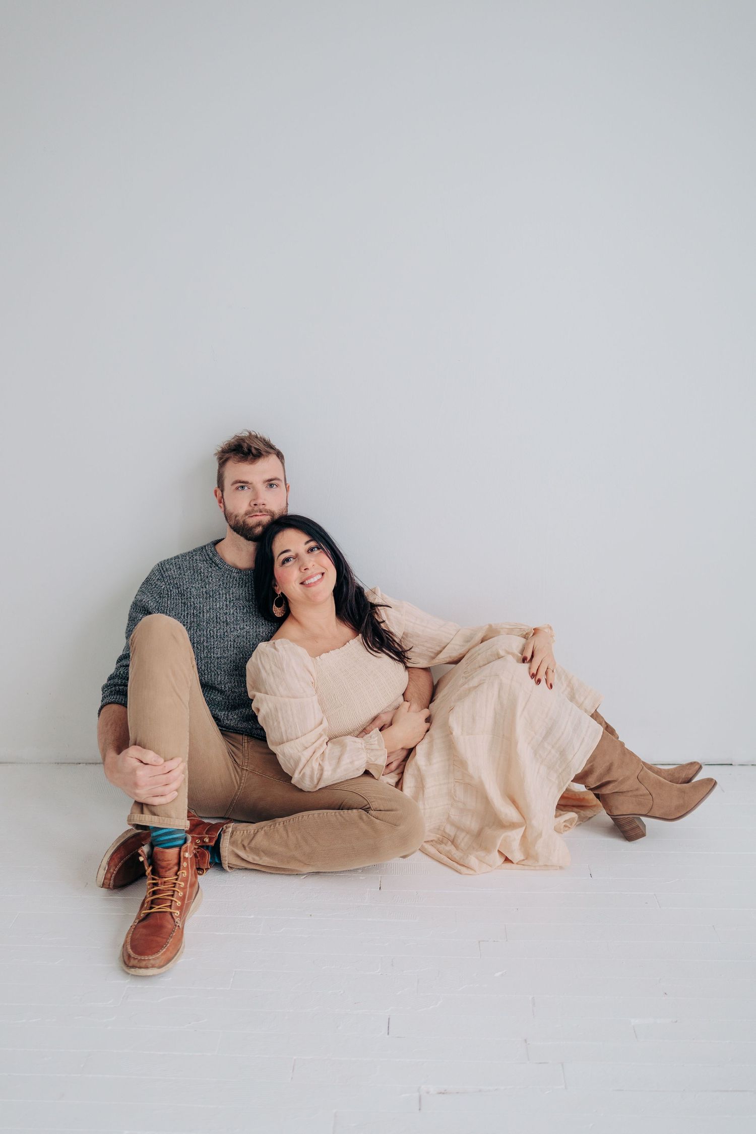 A couple sits together on a white floor wearing casual clothing in soft neutral tones for a minimalist portrait session.