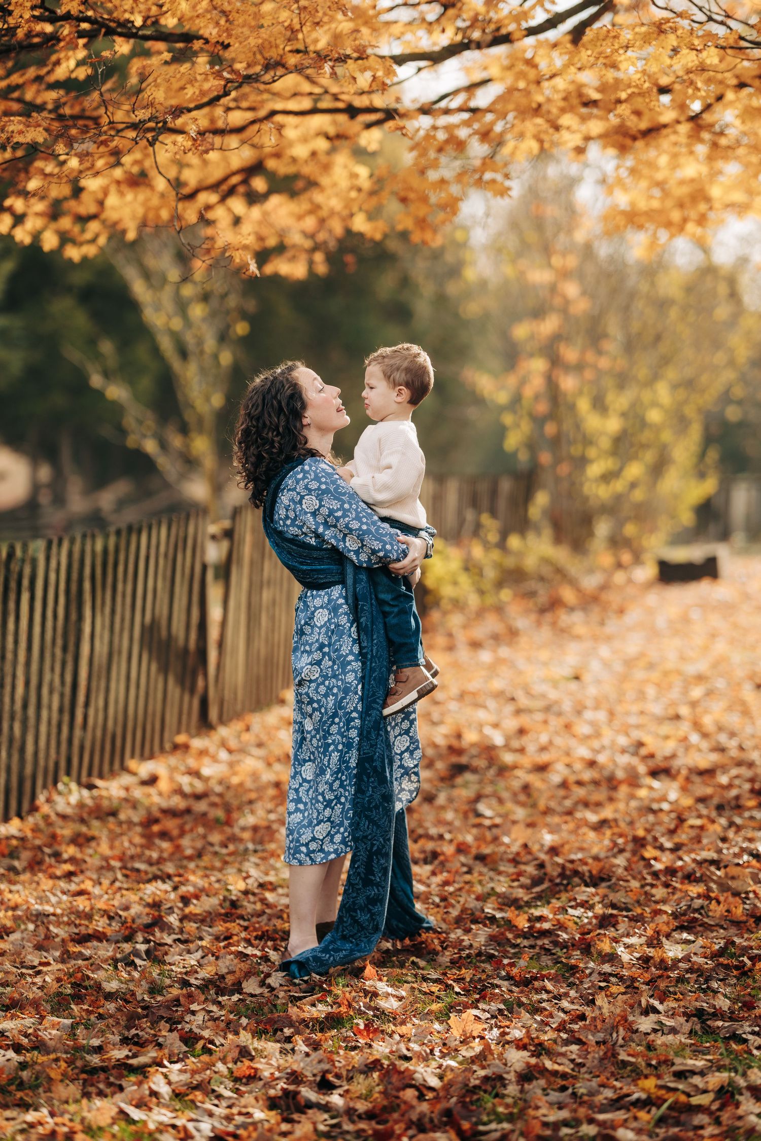 Autumn scene in park with fallen orange leaves and wooden fence as figures embrace among seasonal foliage.