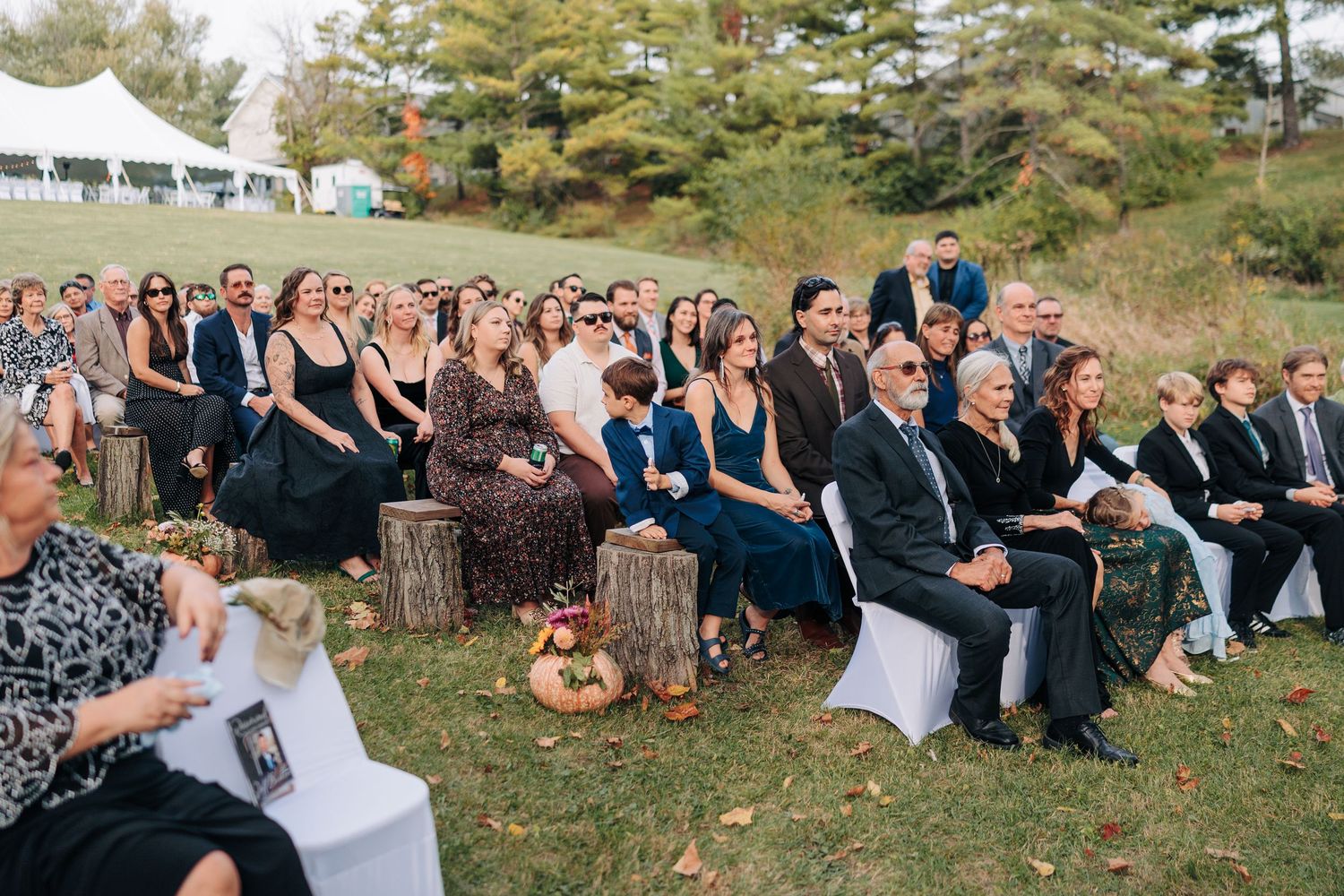 Wedding guests seated outdoors on white chairs during an autumn ceremony.