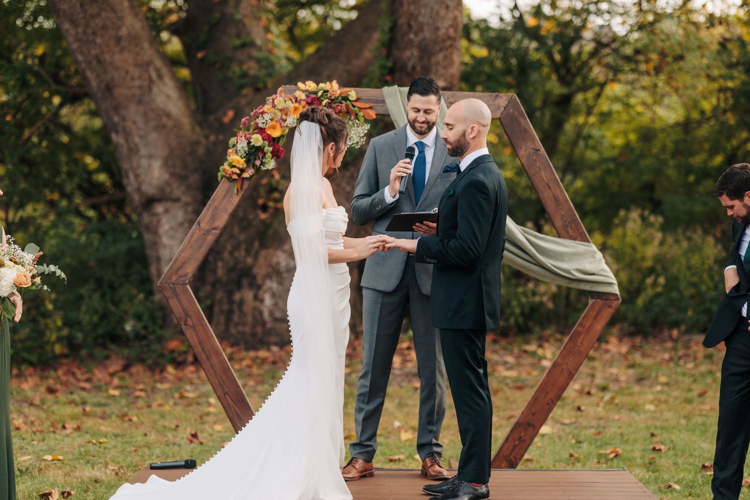 A couple exchanges rings during an outdoor wedding ceremony beneath a rustic wooden arch at sunset.