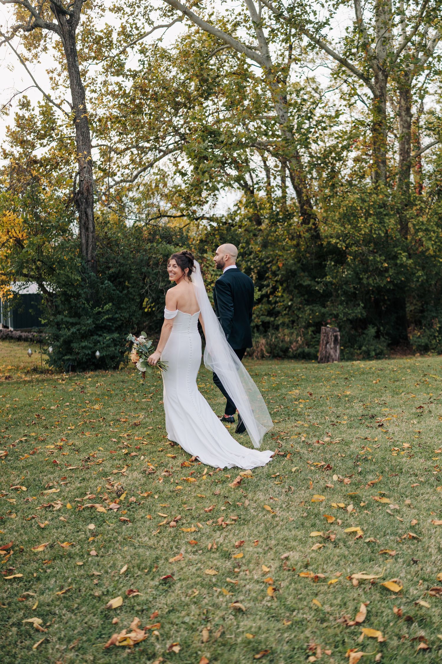 Newlywed couple dancing and twirling in a grassy autumn field with fallen leaves and trees in the background.