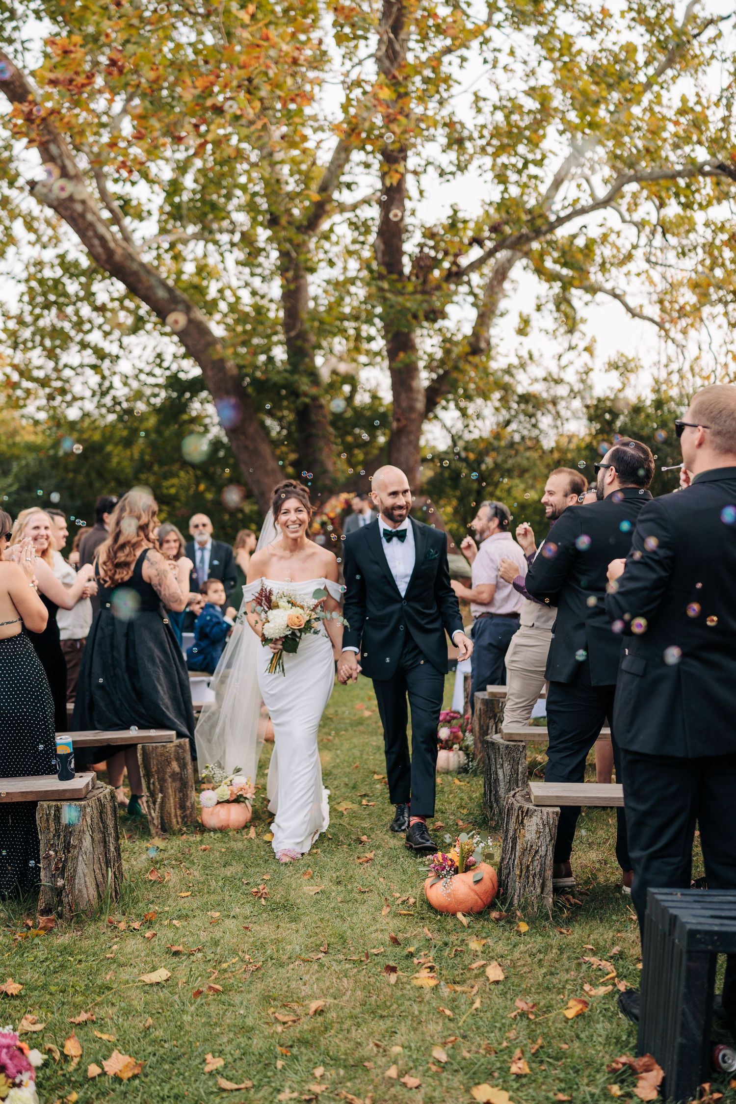 Outdoor autumn wedding ceremony with guests seated on wooden benches surrounded by fall foliage.