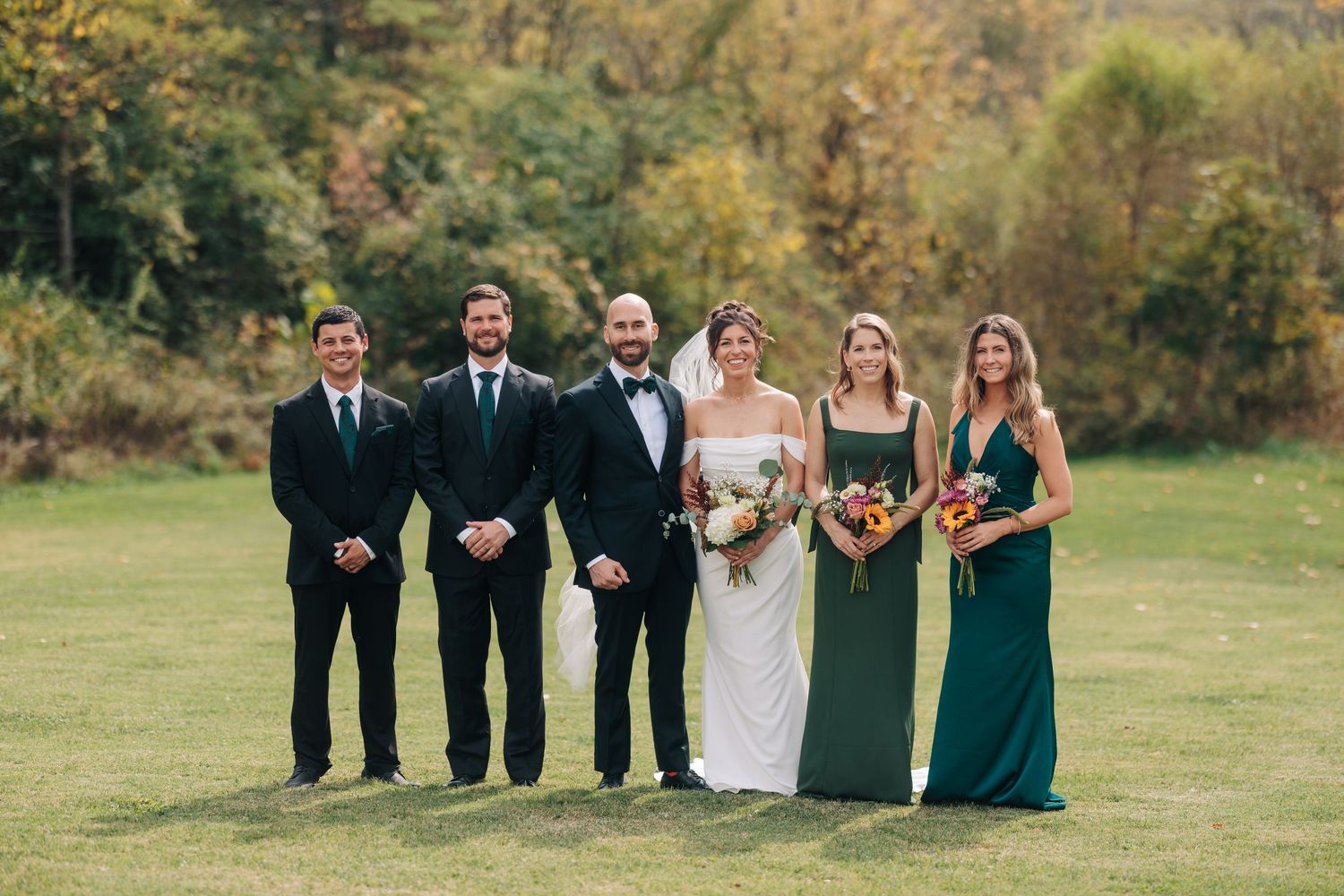 Wedding party stands in a line on green grass with autumn trees in the background wearing formal attire.