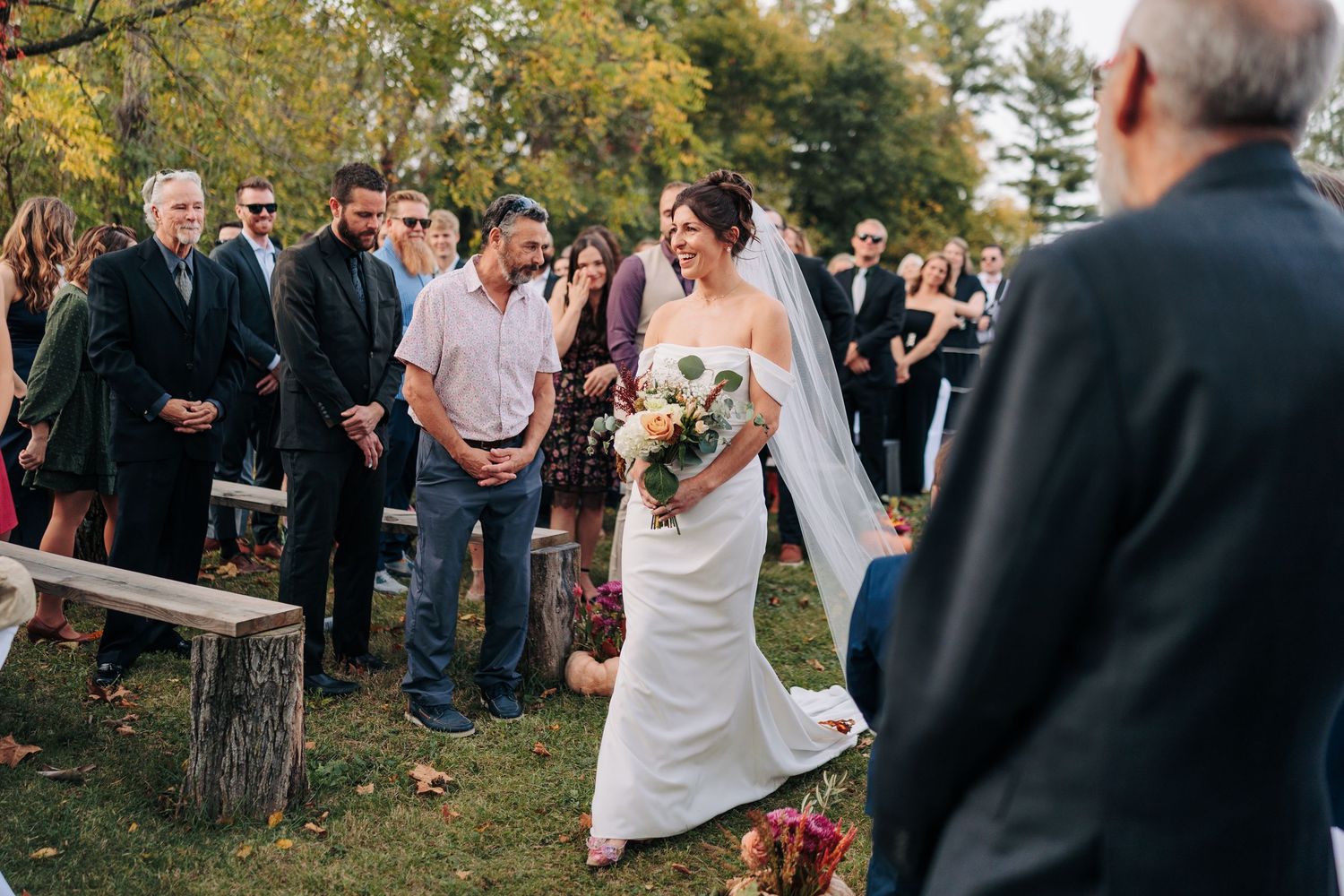 Wedding guests gather outdoors in a natural setting during a fall celebration with trees in the background.