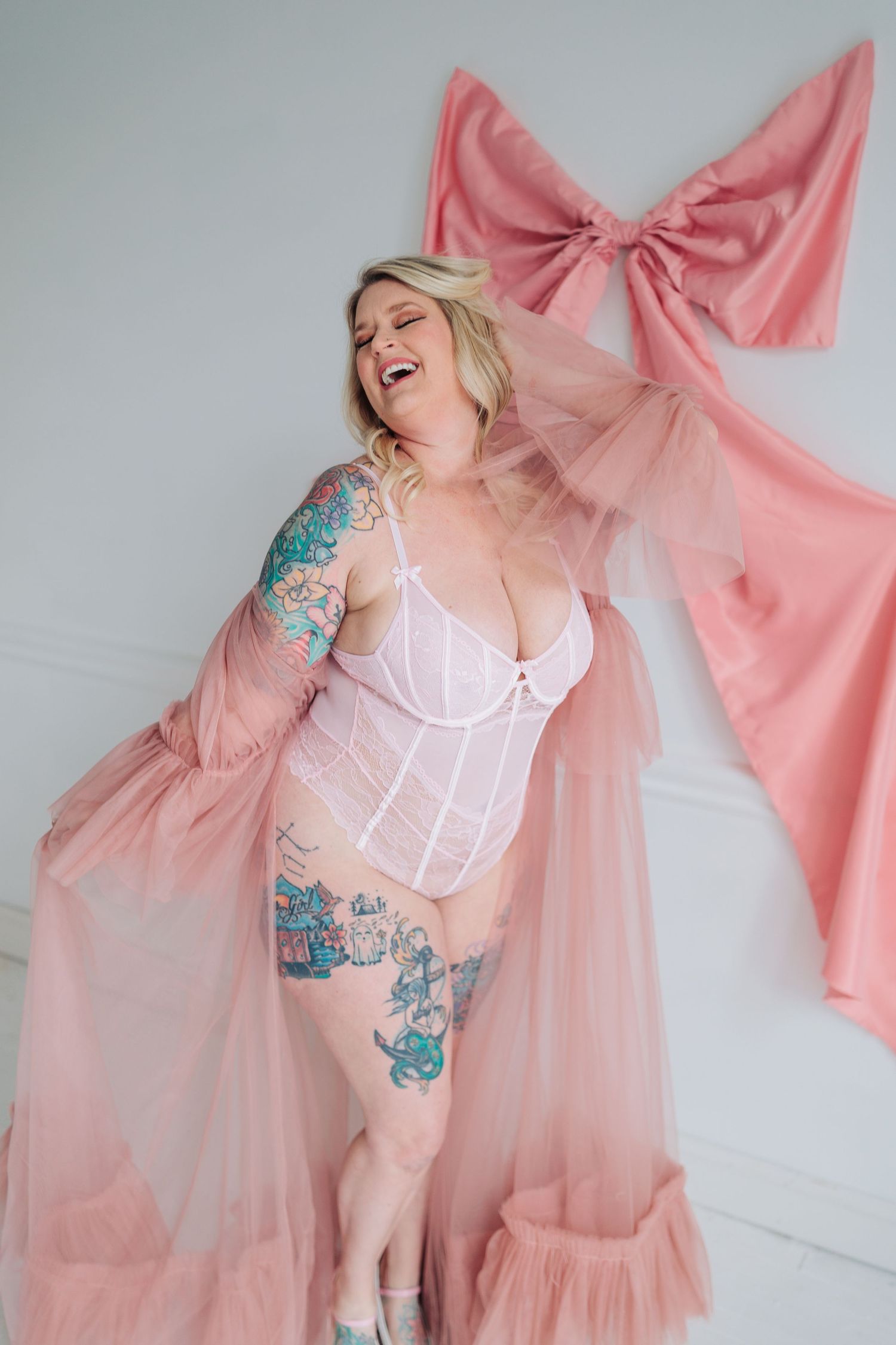 Person wearing white lingerie and pink sheer robe poses joyfully with colorful tattoos against pink bow backdrop.