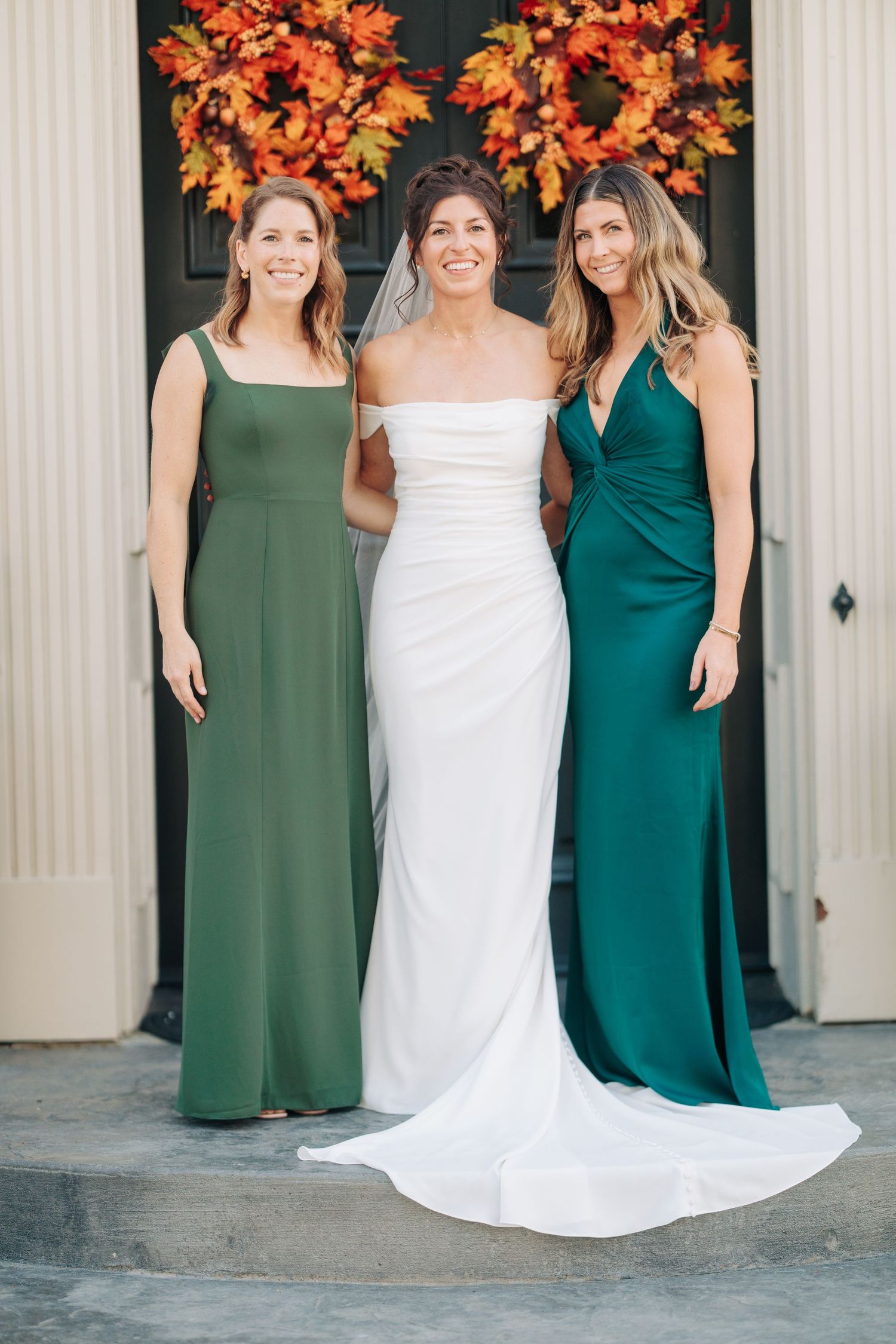 Wedding party poses in front of orange fall wreaths wearing elegant green and white dresses.