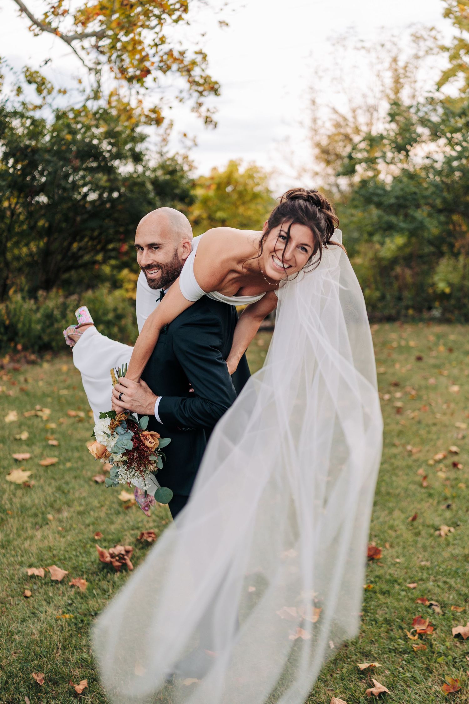A bride in a flowing white dress spins playfully in an autumn garden with fallen leaves on the ground.