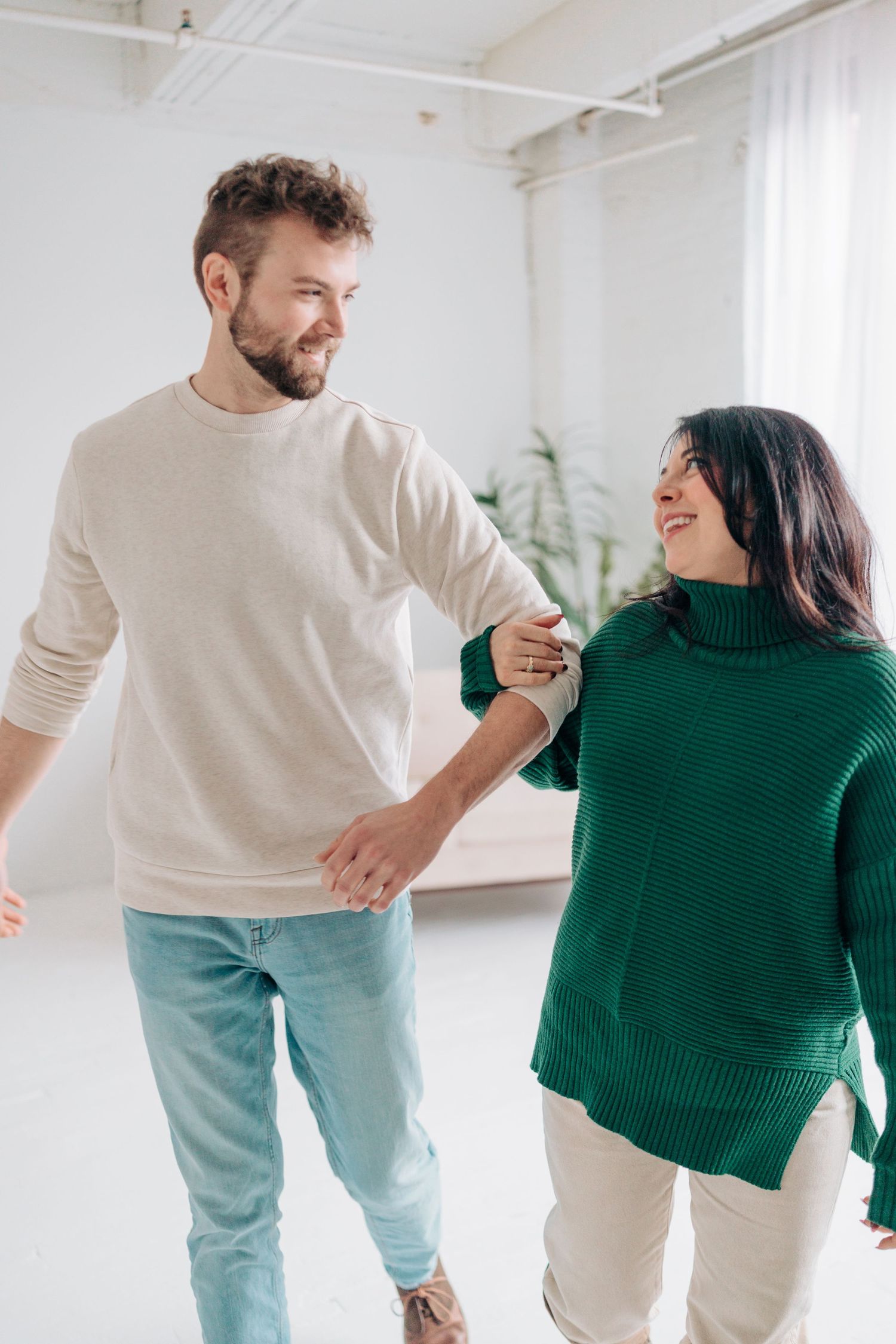 A couple in light blue jeans and contrasting sweaters dance playfully together in a bright white studio space.