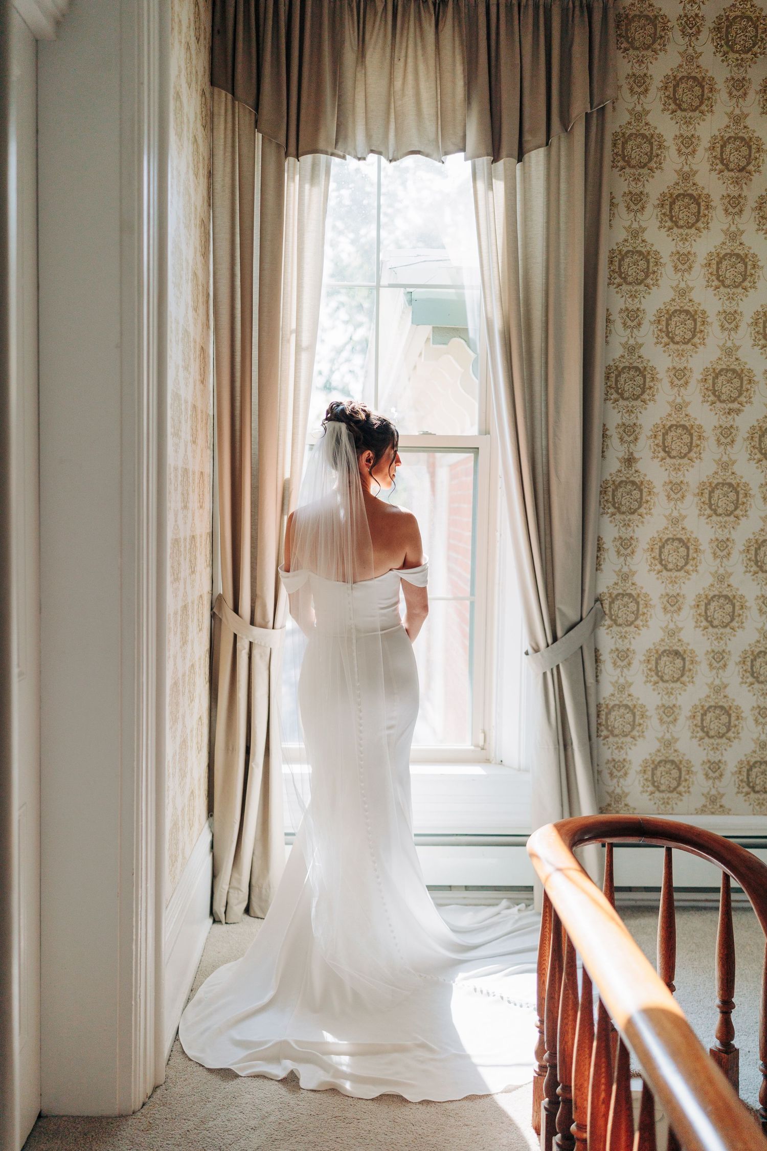 A bride in a flowing white dress gazes out a tall window with vintage wallpaper and curtains in natural light.