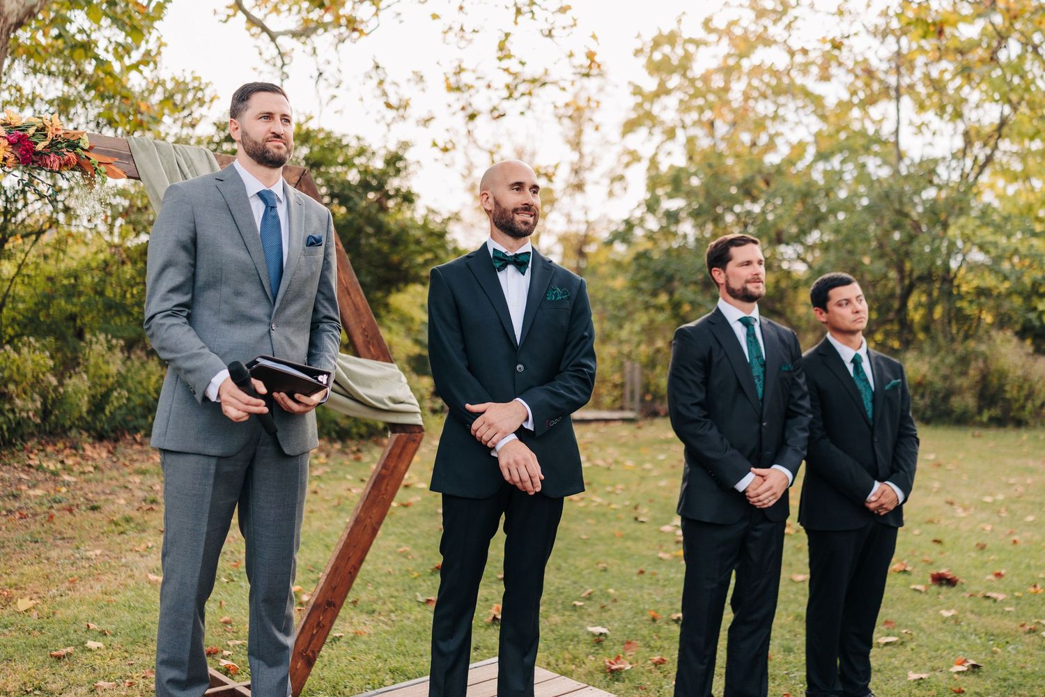 Wedding party members in gray suits stand together on grass during an autumn outdoor ceremony.