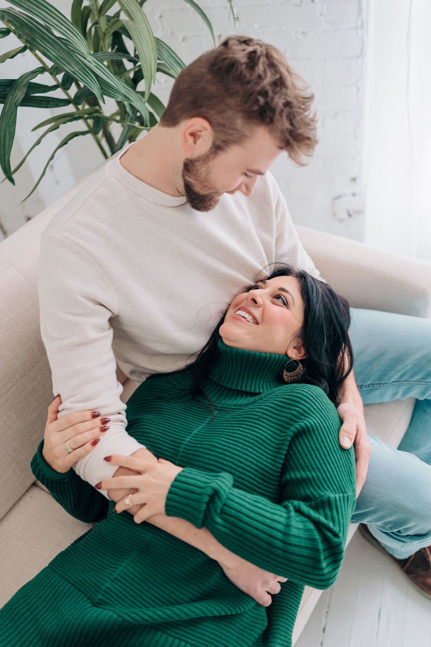 A couple in green and beige clothing embraces on a light colored sofa in a bright minimalist setting.