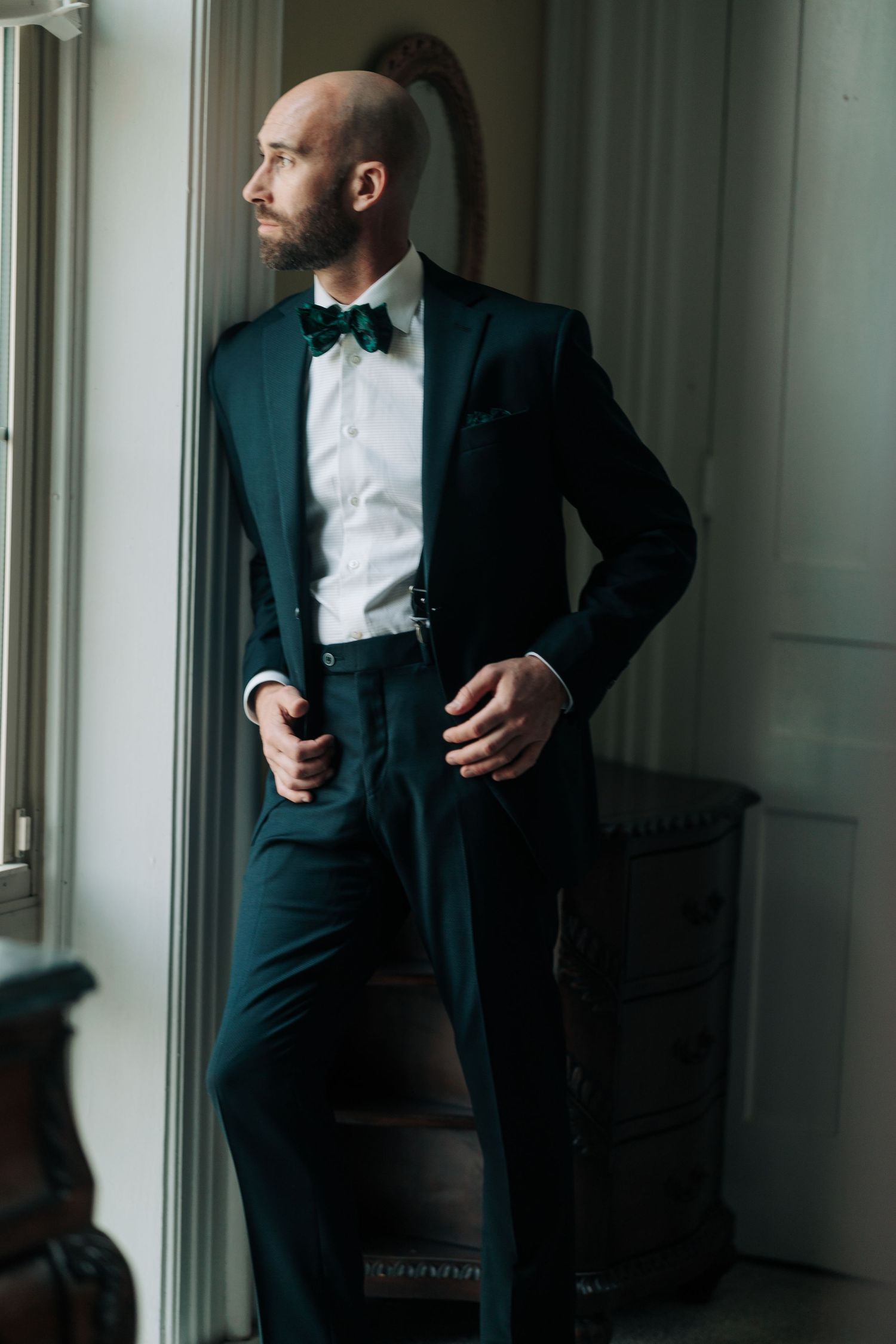 Groom in classic black tuxedo adjusts his suit while standing by window light.