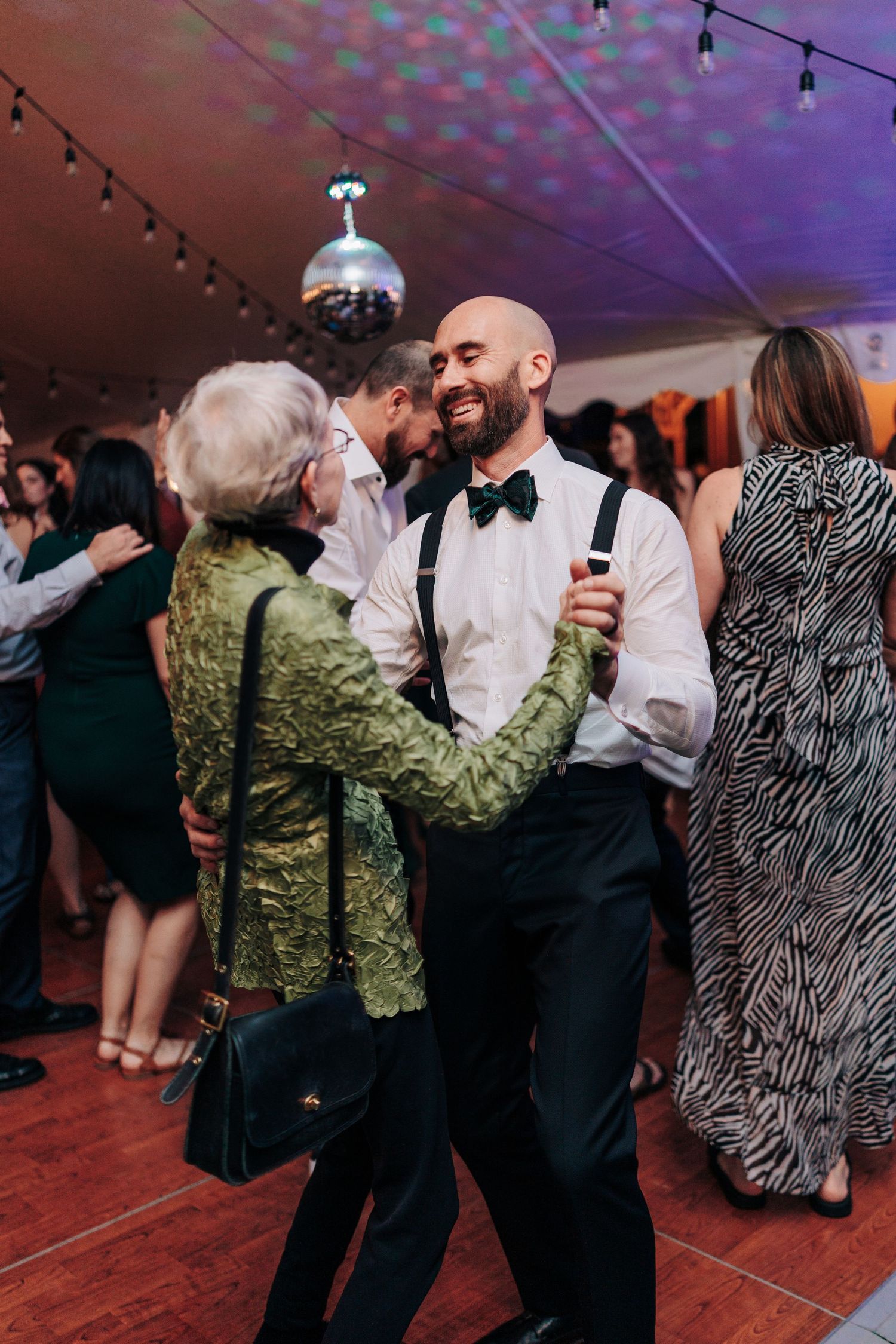 Wedding guests dance together in formal attire under purple lighting at an evening reception celebration.
