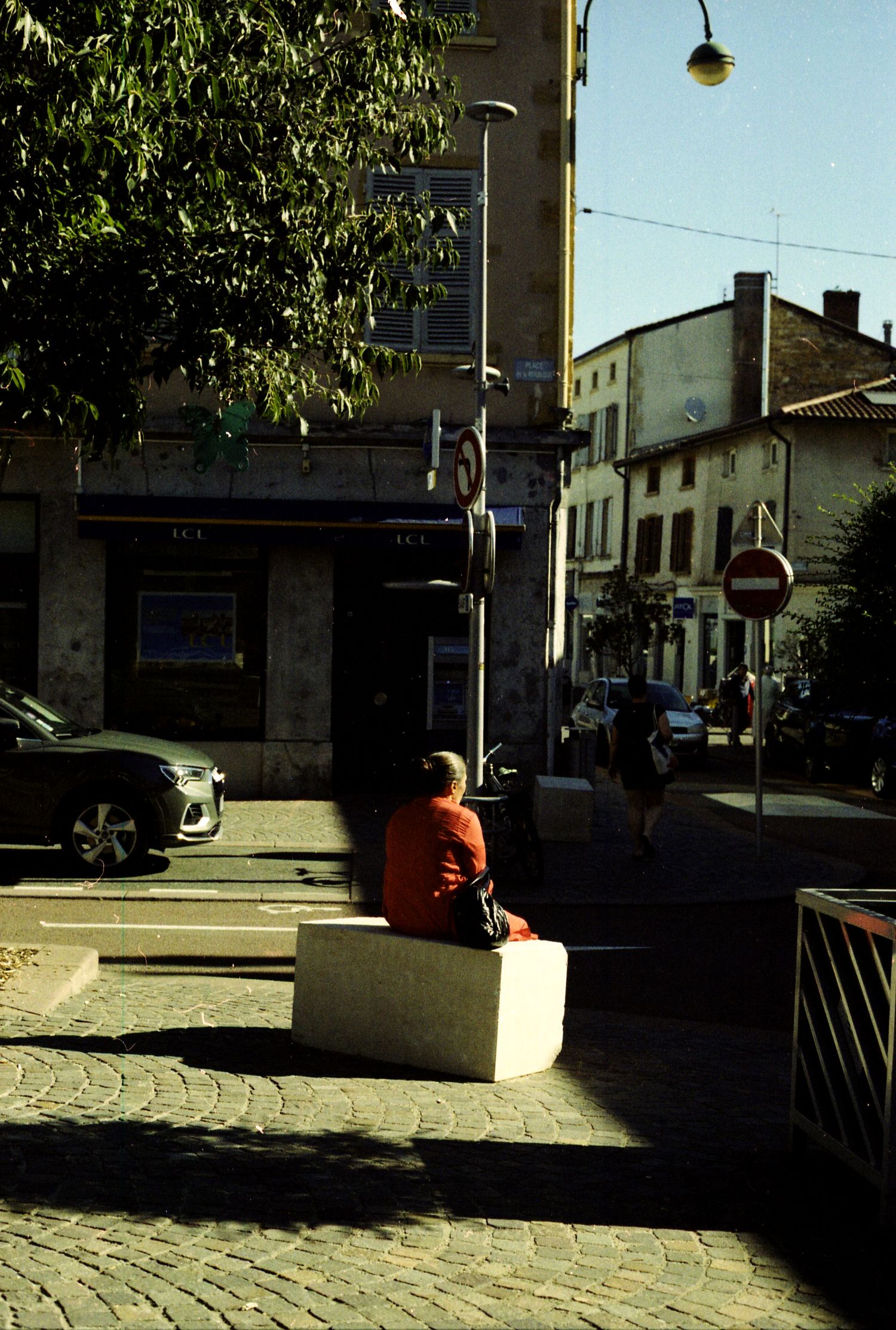Photo  de rue argentique. La lumière est contrastée, une femme avec un haut rouge est assise de dos sur un banc en béton