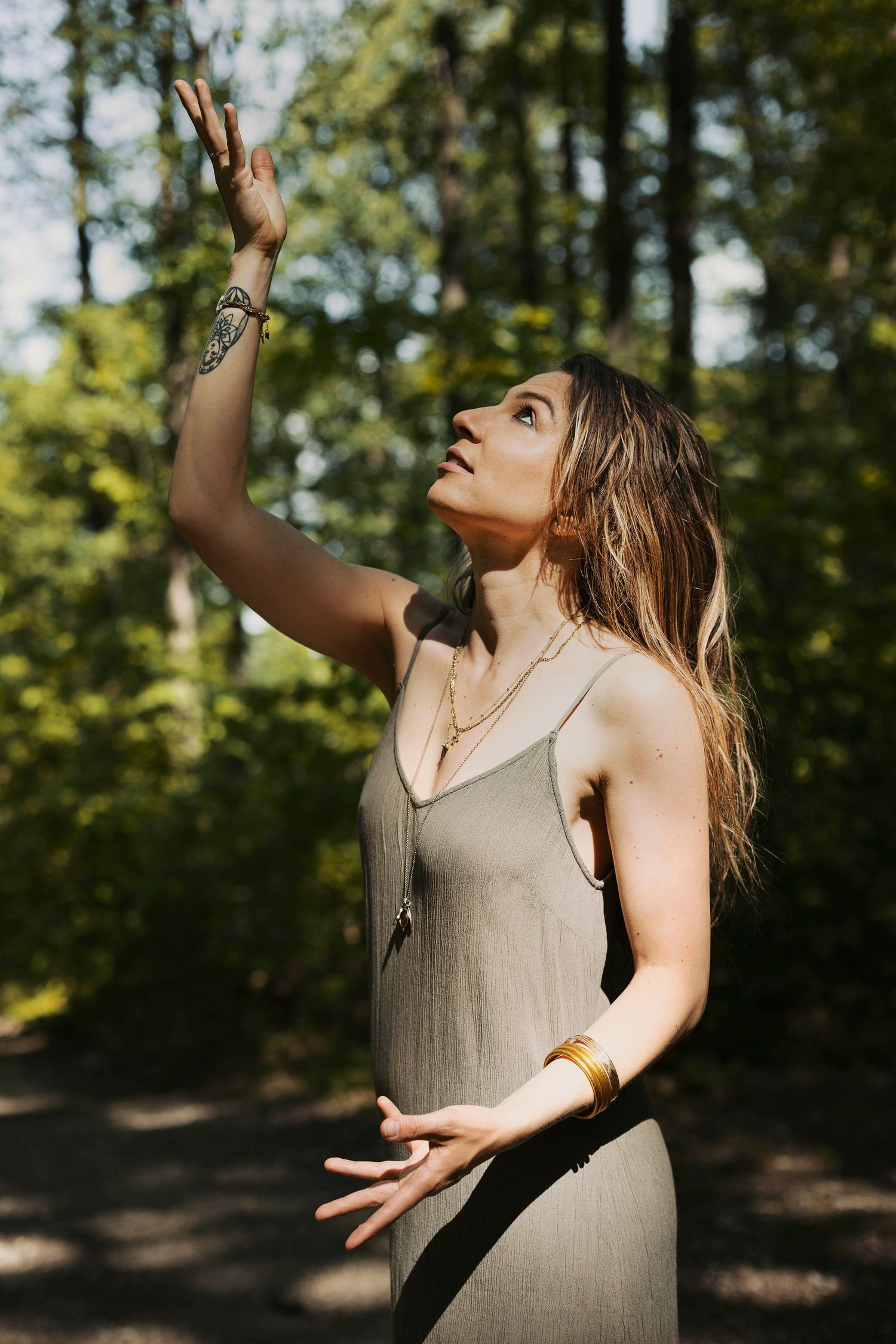 Portrait d'une femme, main et regard tournés vers le ciel, dans une robe fluide verte, arrière plan forestier.