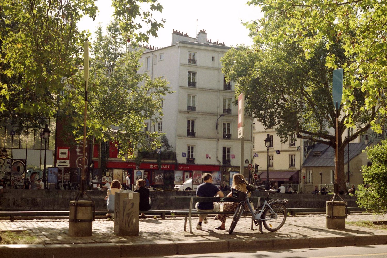 Photo de rue argentique avec des personnes assises sur un banc, une bicyclette est garée à proximité. Immeuble parisien en arrière plan.