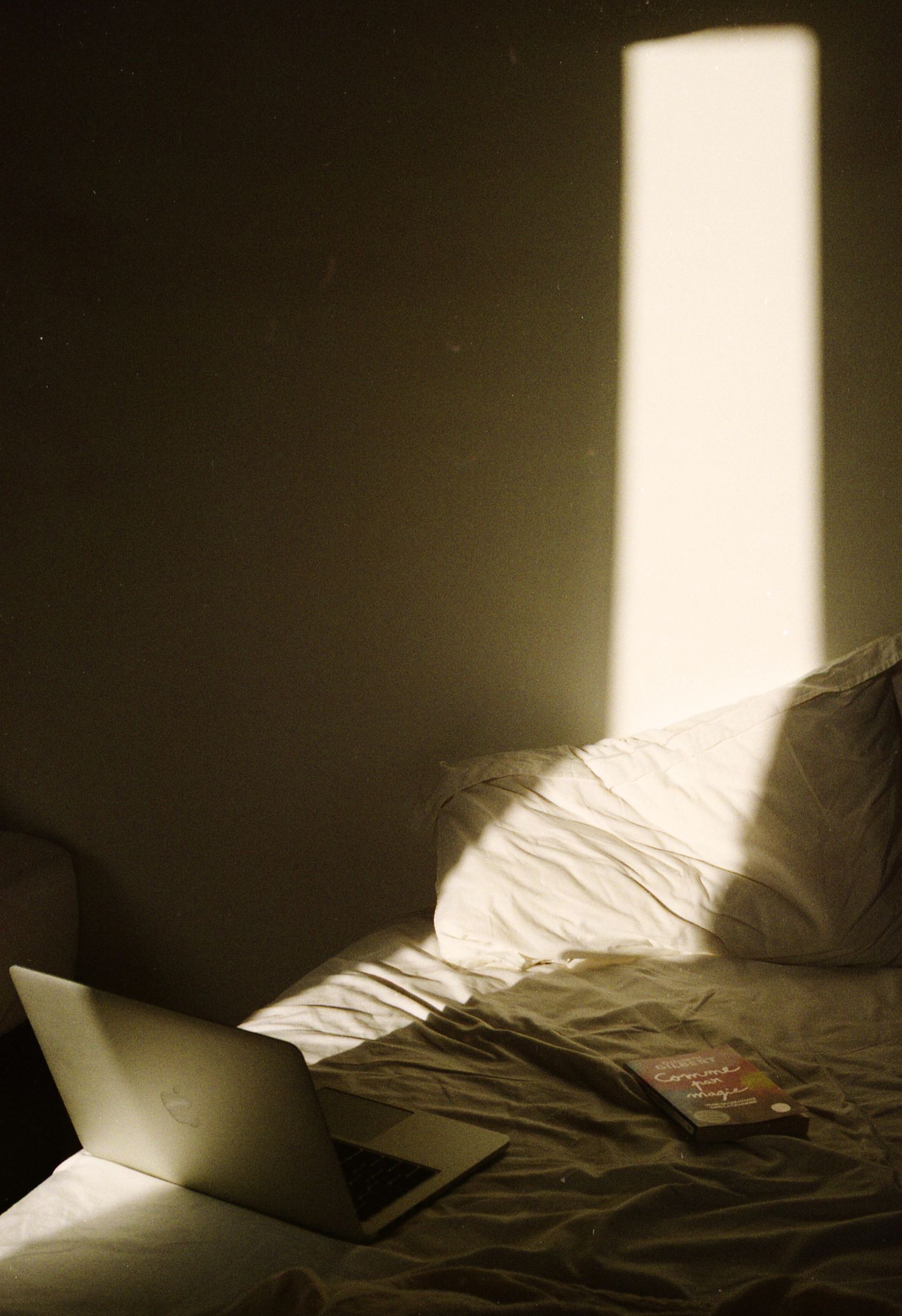 Photo argentique d'un mac et d'un livre posés sur un lit, illuminés par un rectangle de lumière.