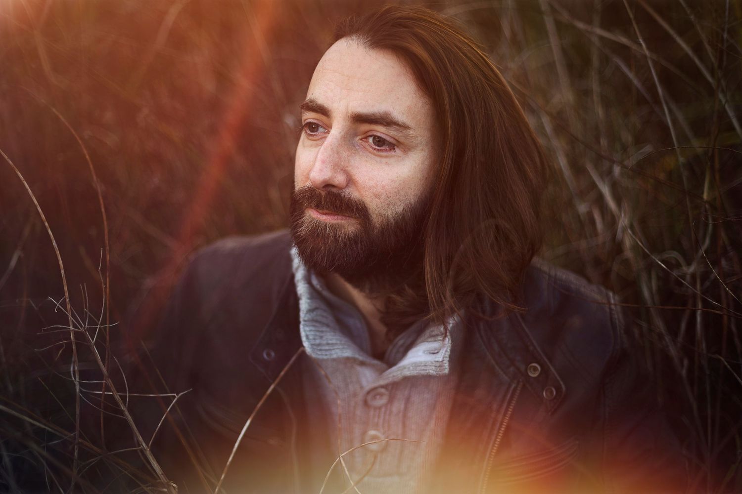 Portrait d'un homme brun aux cheveux long, regardant ailleurs, entouré d'herbes hautes.