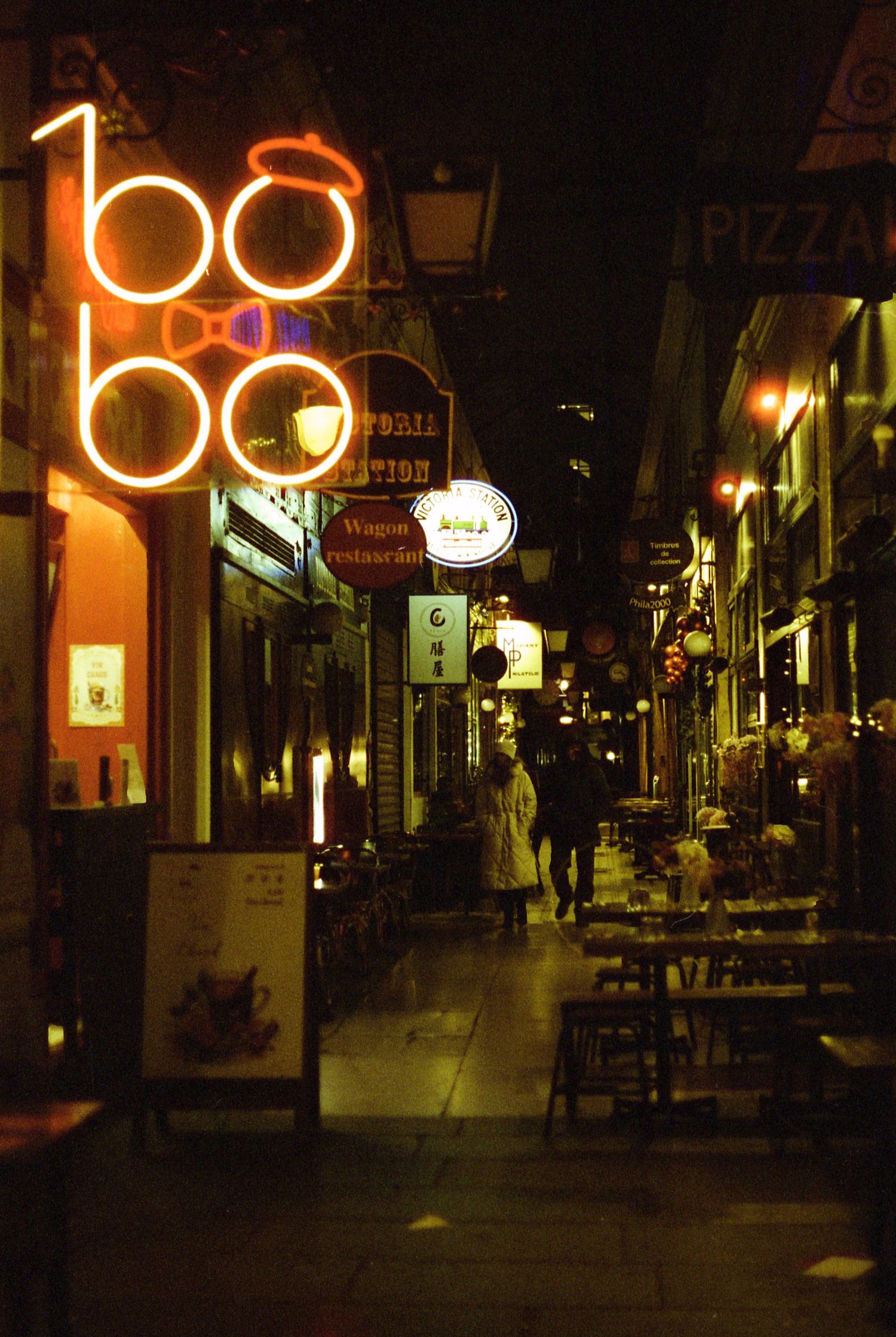 Photo de rue argentique du passage des panoramas, la nuit. Les néons des enseignes illuminent ce passage parisien