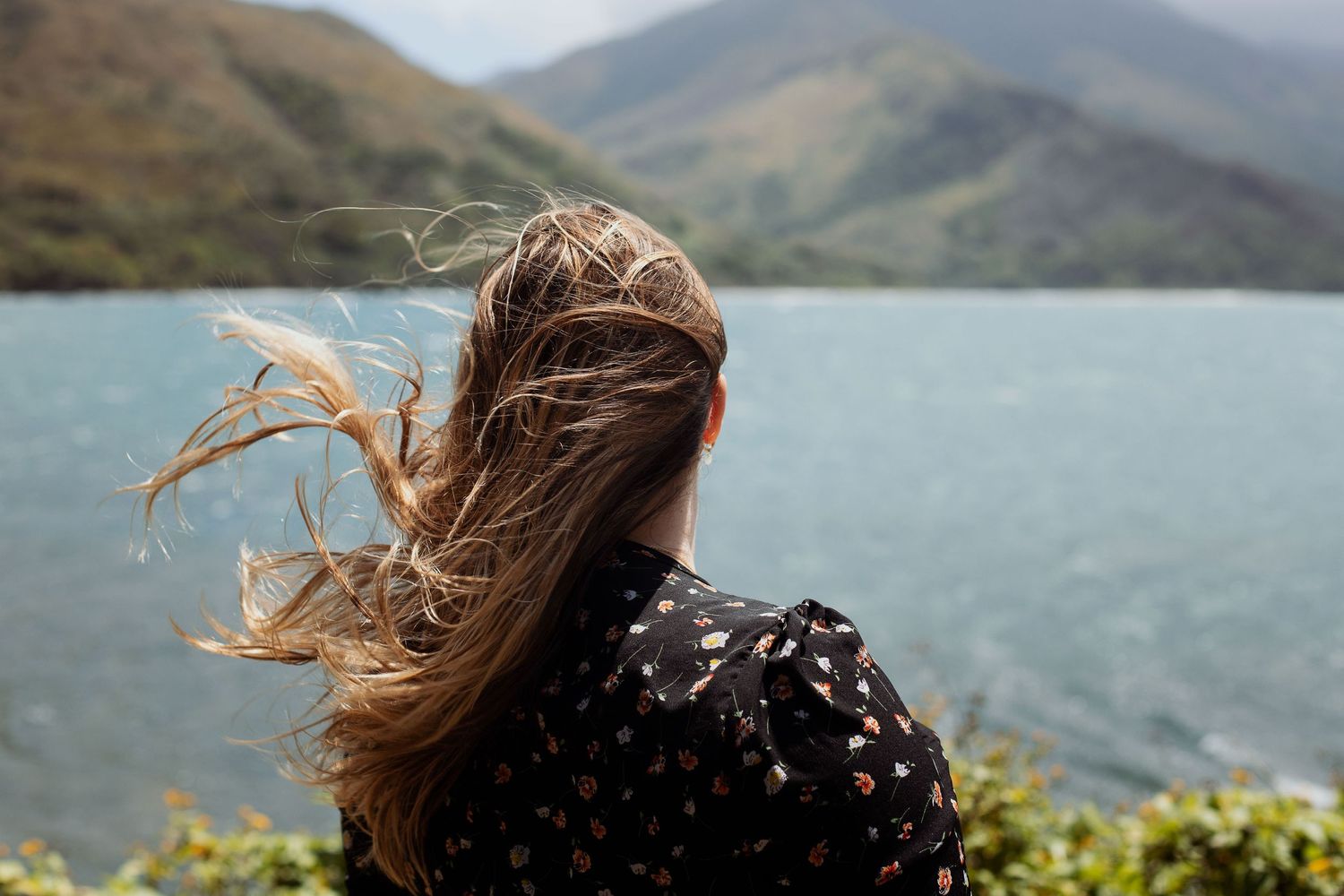 Photo d'une femme blonde cheveux aux vent face à une étendue d'eau et des montagnes en arrière plan