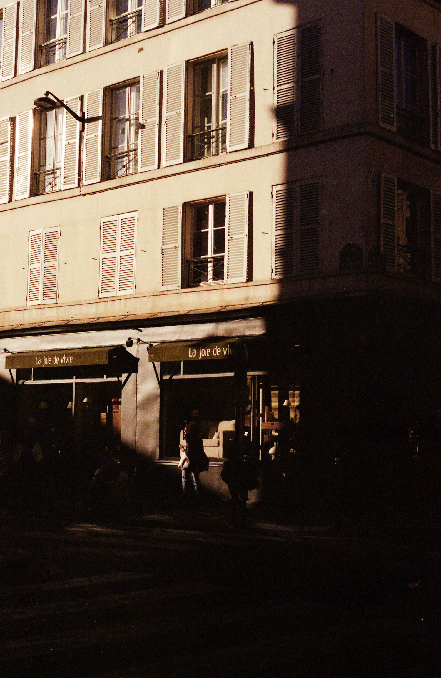 Photo de rue argentique d'un immeuble parisien à moitié illuminé par le soleil. Nous pouvons y lire sur la devanture : joie de vivre