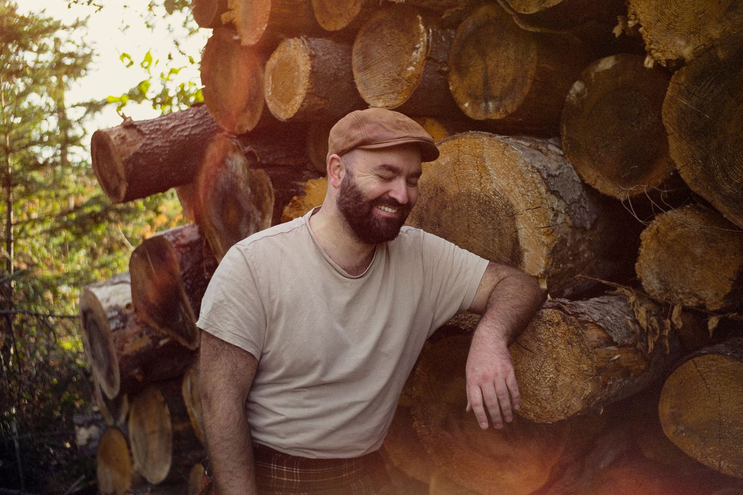 Portrait d'un homme souriant posant accoudé à des tronc d'arbre empilés.