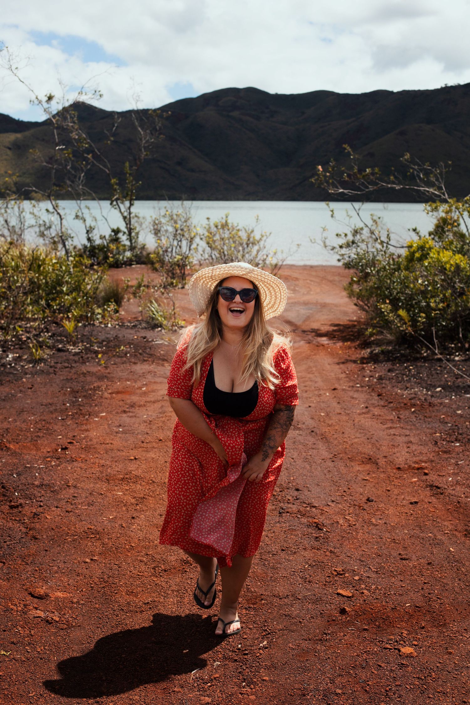 Photo d'une femme souriante devant un paysage naturel composé de terre rouge sur le sol, lac et montagnes en arrière plan