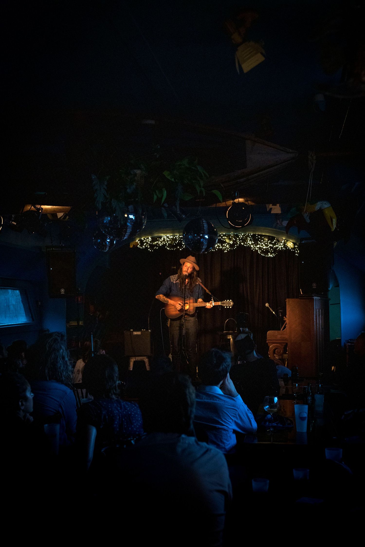Photo de concert, on y voit le public dans la pénombre, et l'artiste sur scène éclairé en orange avec sa guitare