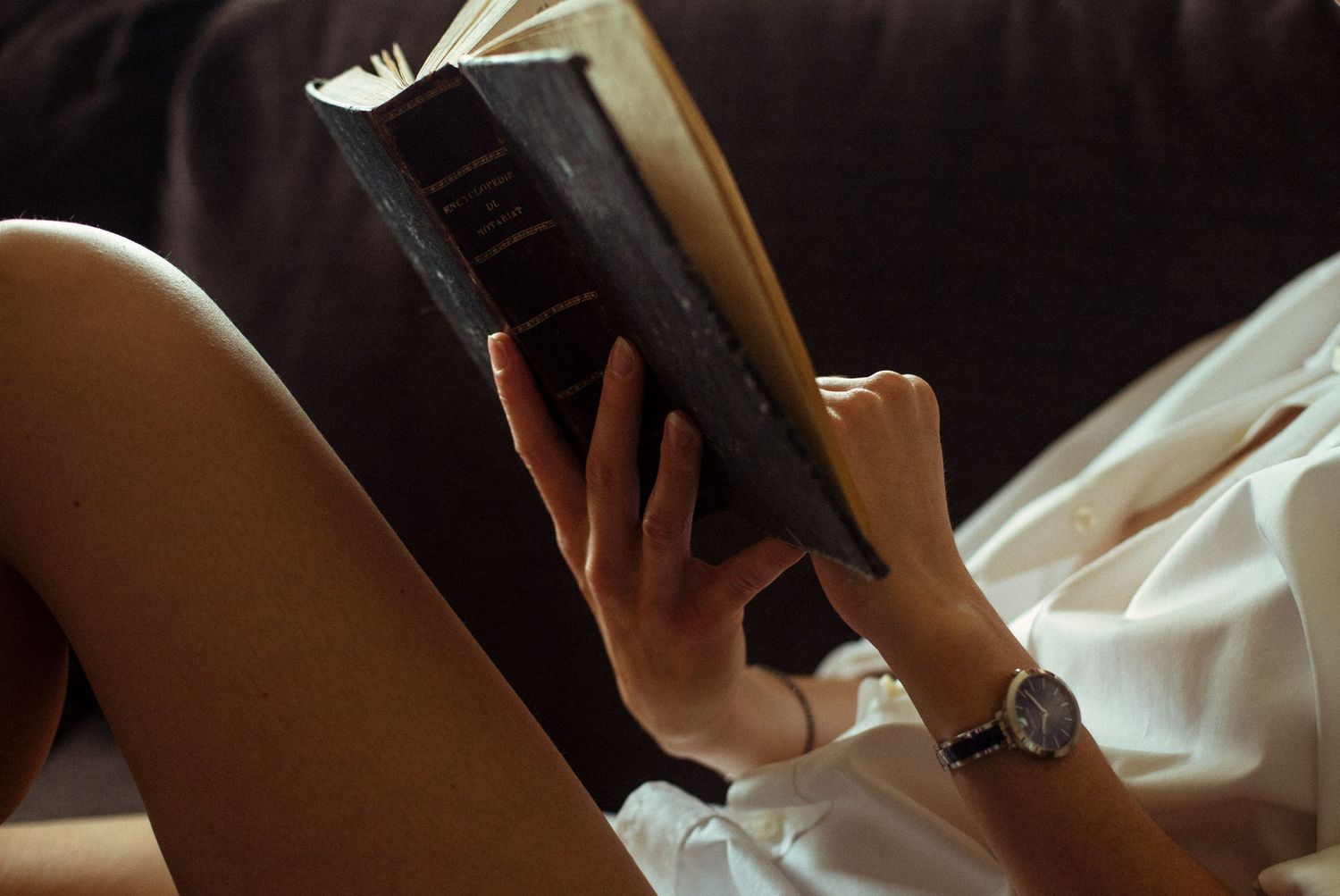 Photo détail intimiste d'une femme lisant un livre, en chemise blanche et jambes nues