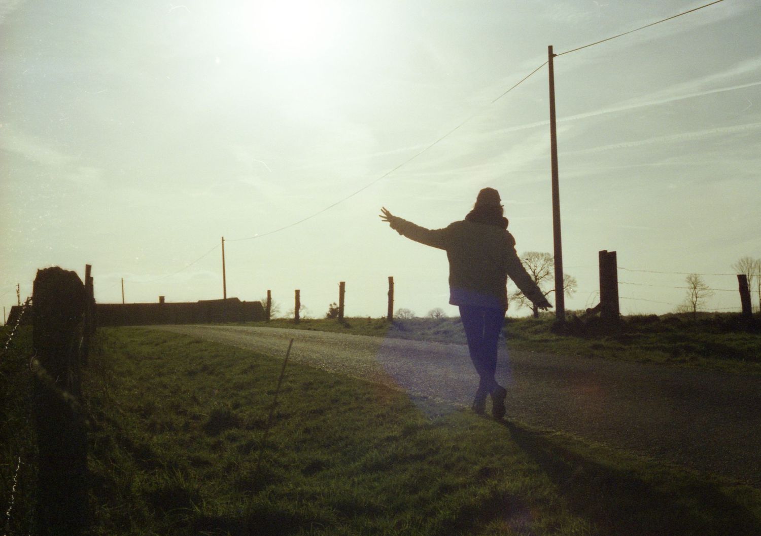 Autoportrait à l'argentique d'une femme  de dos marchant sur une route de campagne entourée de champs