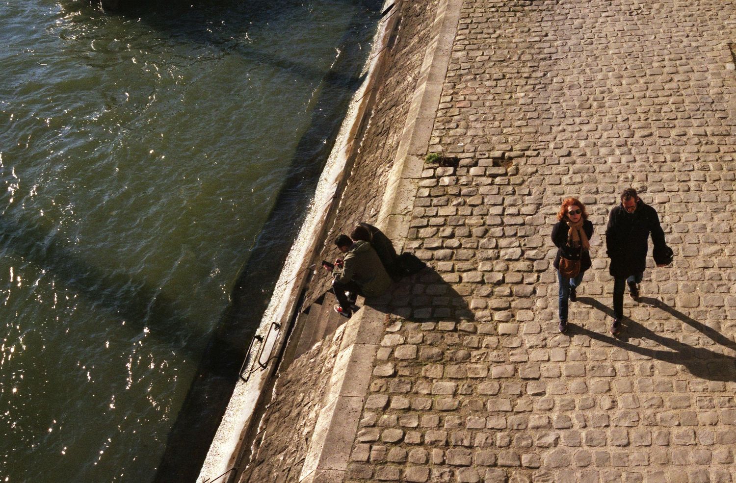 Photo de rue argentique prise en hauteur. En contrebas, couple est assis sur les berges de seine, tandis qu'un autre, au même niveau marche.