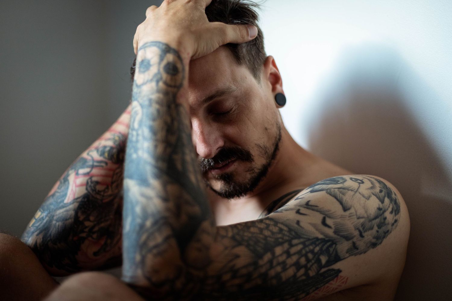 Portrait intimiste d'un homme torse nu, les bras tatoués et se prenant le visage dans les mains, sur fond neutre.