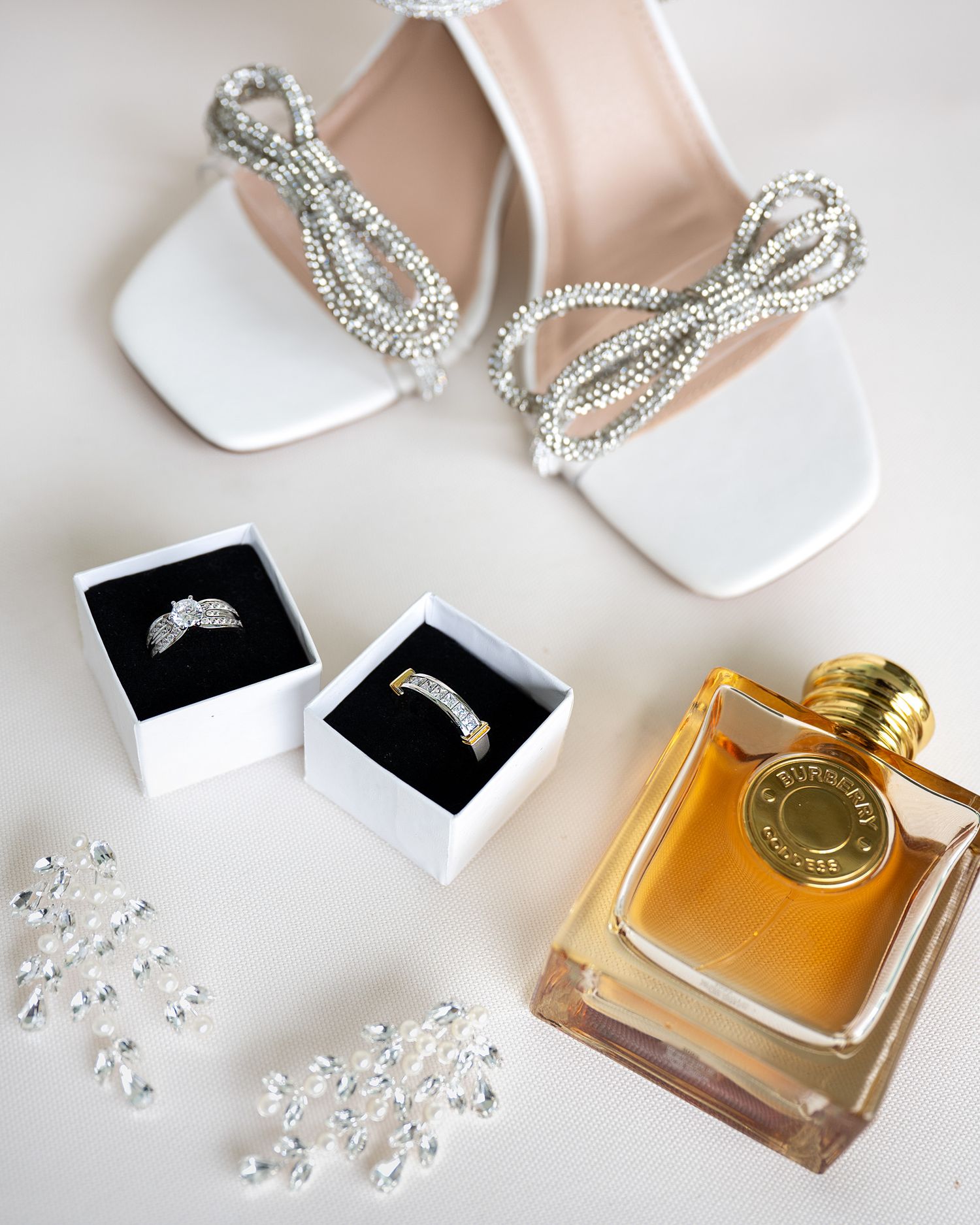 Bridal flat lay with white heeled sandals, two wedding rings in boxes, crystal earrings, and amber perfume bottle.