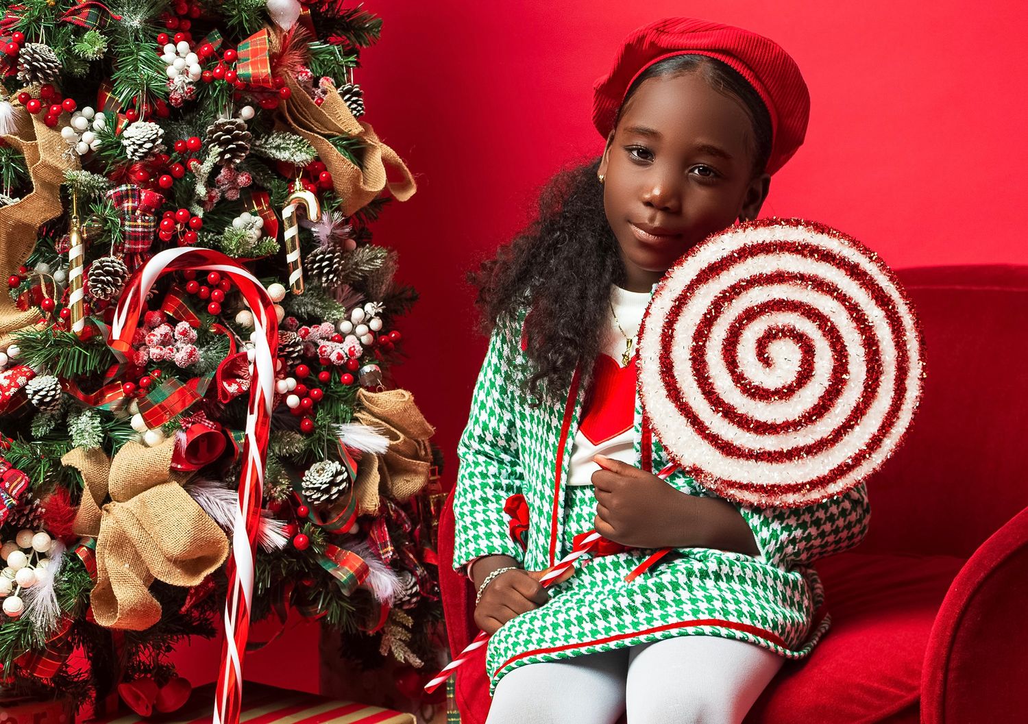 A festive holiday scene with a decorated Christmas tree and large candy cane prop against a red backdrop.