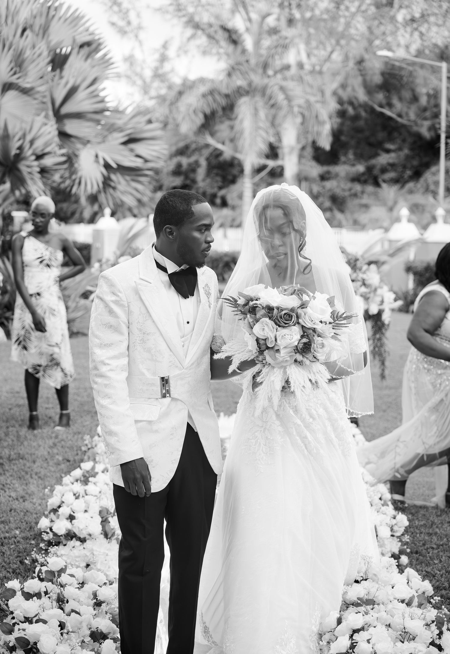Black and white wedding ceremony photo with couple walking down a flower-lined aisle outdoors.