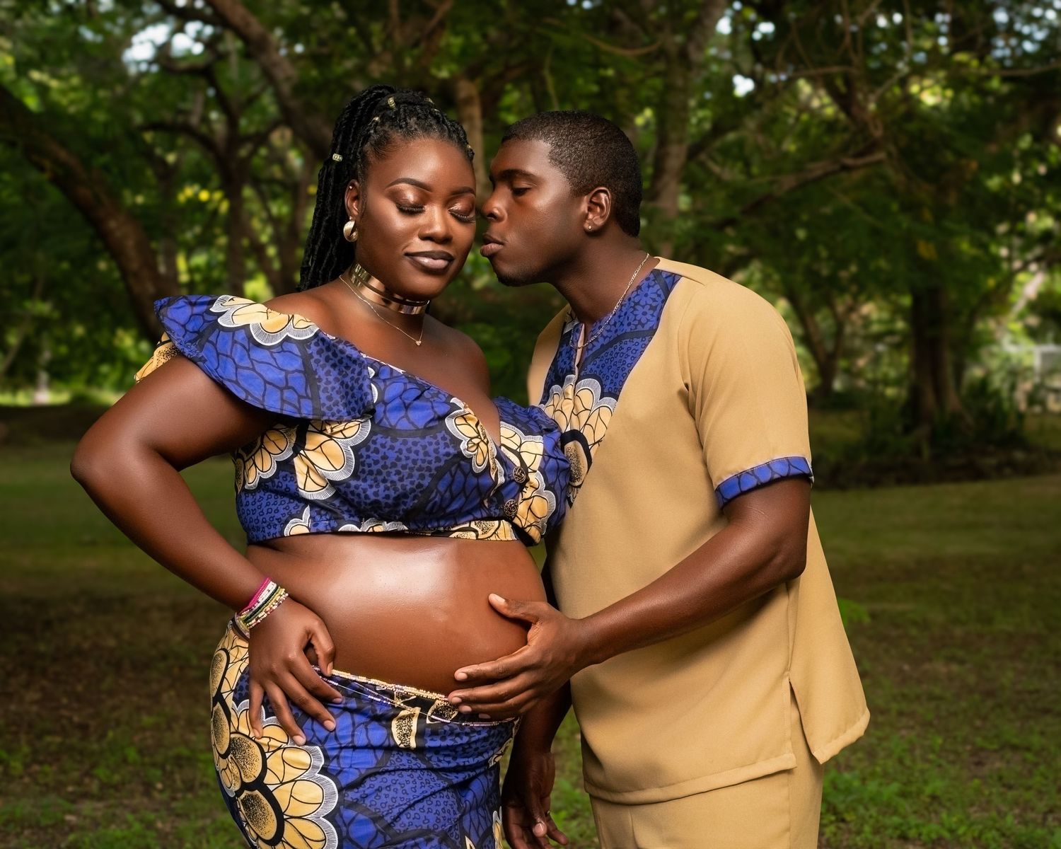 A maternity portrait of a couple in matching blue and gold African print outfits in a garden setting.