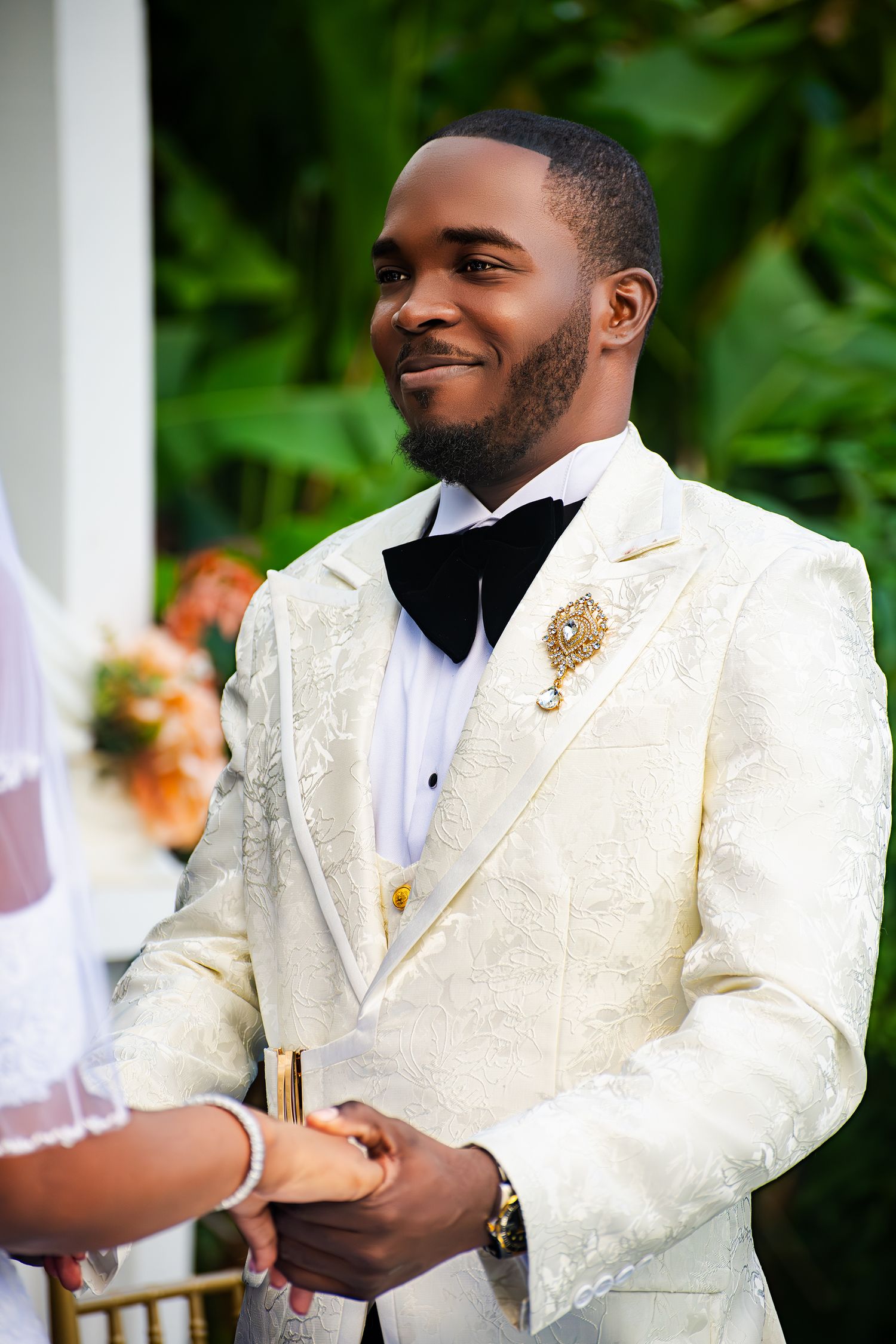 Groom in elegant white brocade tuxedo with black bowtie and gold brooch at outdoor wedding.