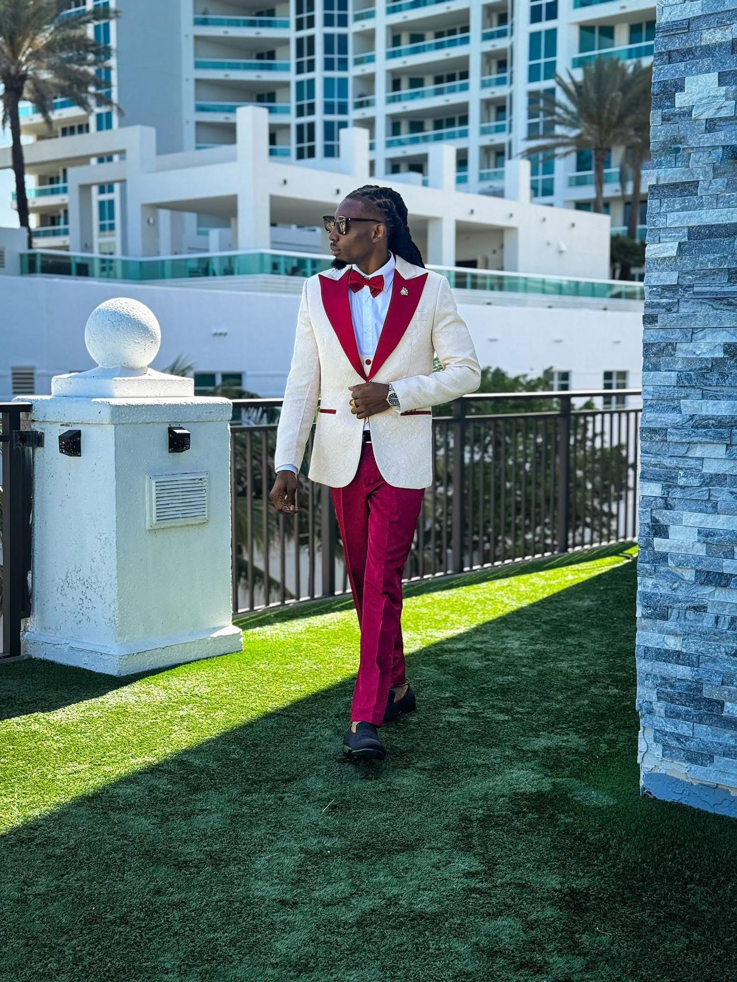 A stylish figure in red pants and white blazer poses confidently on artificial turf near modern buildings and palm trees.