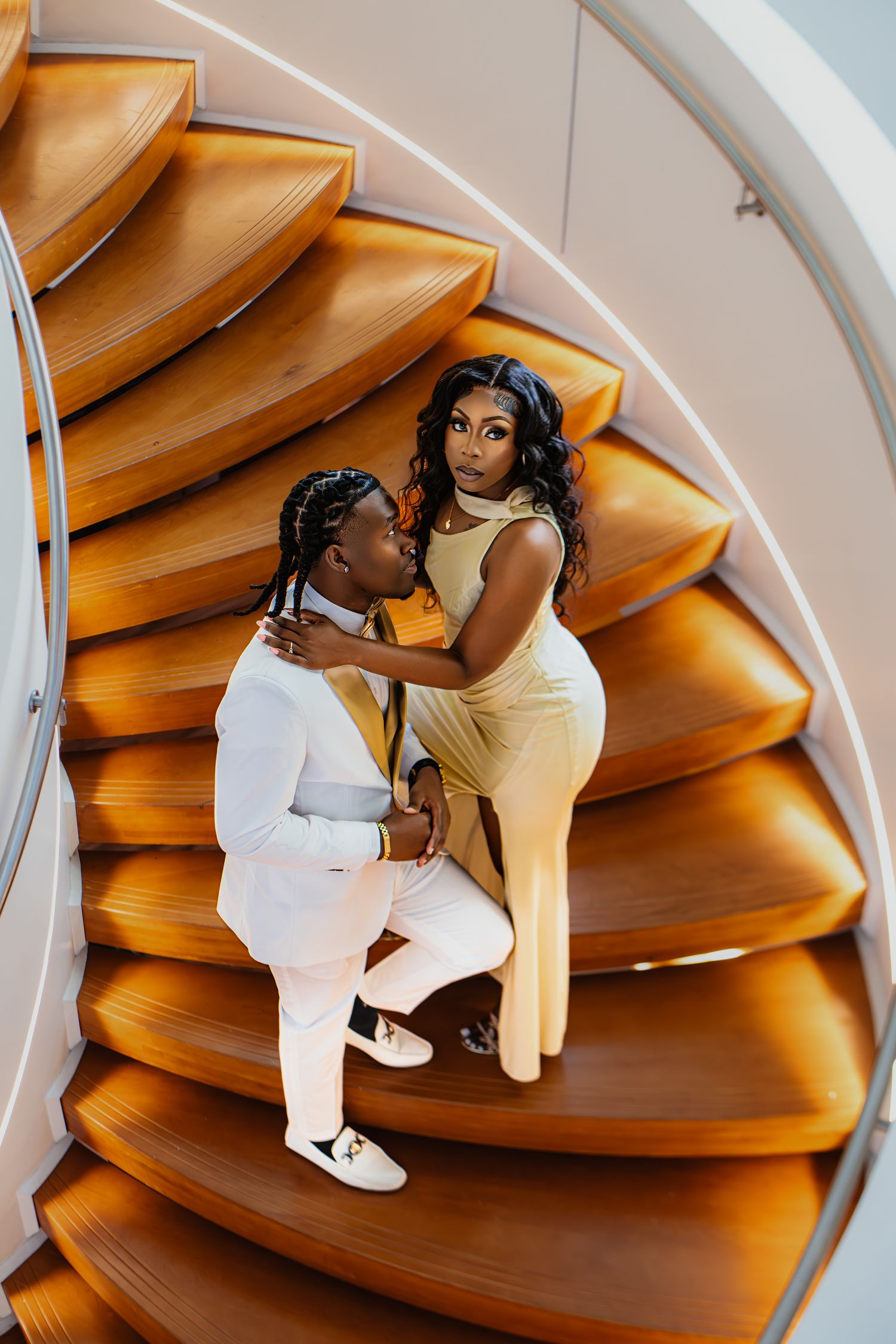 Couple in formal attire pose romantically on curved wooden staircase.