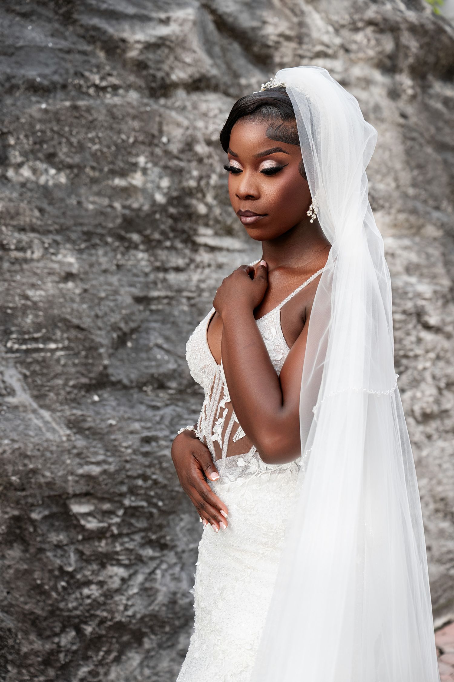 Elegant Black bride in white lace gown and veil poses gracefully against stone wall.
